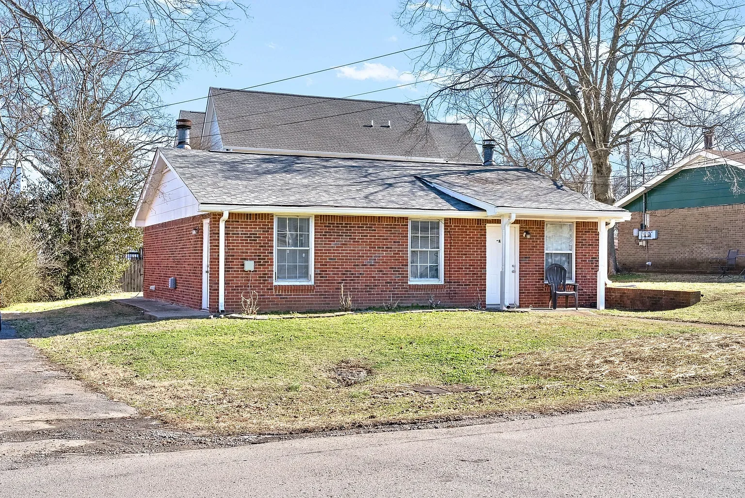 A small brick house with two windows, a white door, a grey shingled roof, and a black chair outside. It is surrounded by a grassy lawn and trees with bare branches in the background.