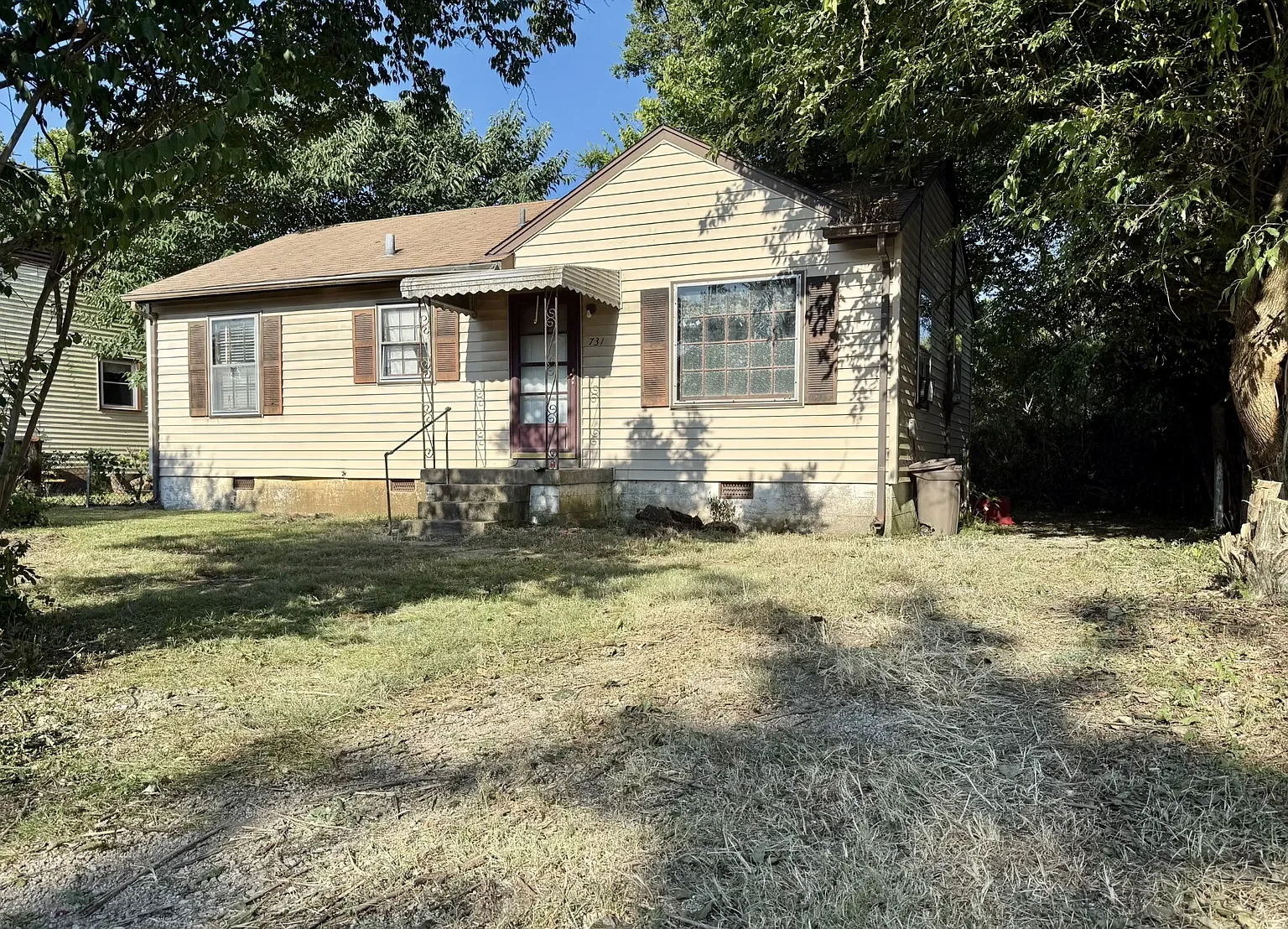A small, single-story house with beige siding, brown shutters, and a front porch with stone steps. The house is surrounded by a yard with grass and trees, and a blue sky is visible overhead.