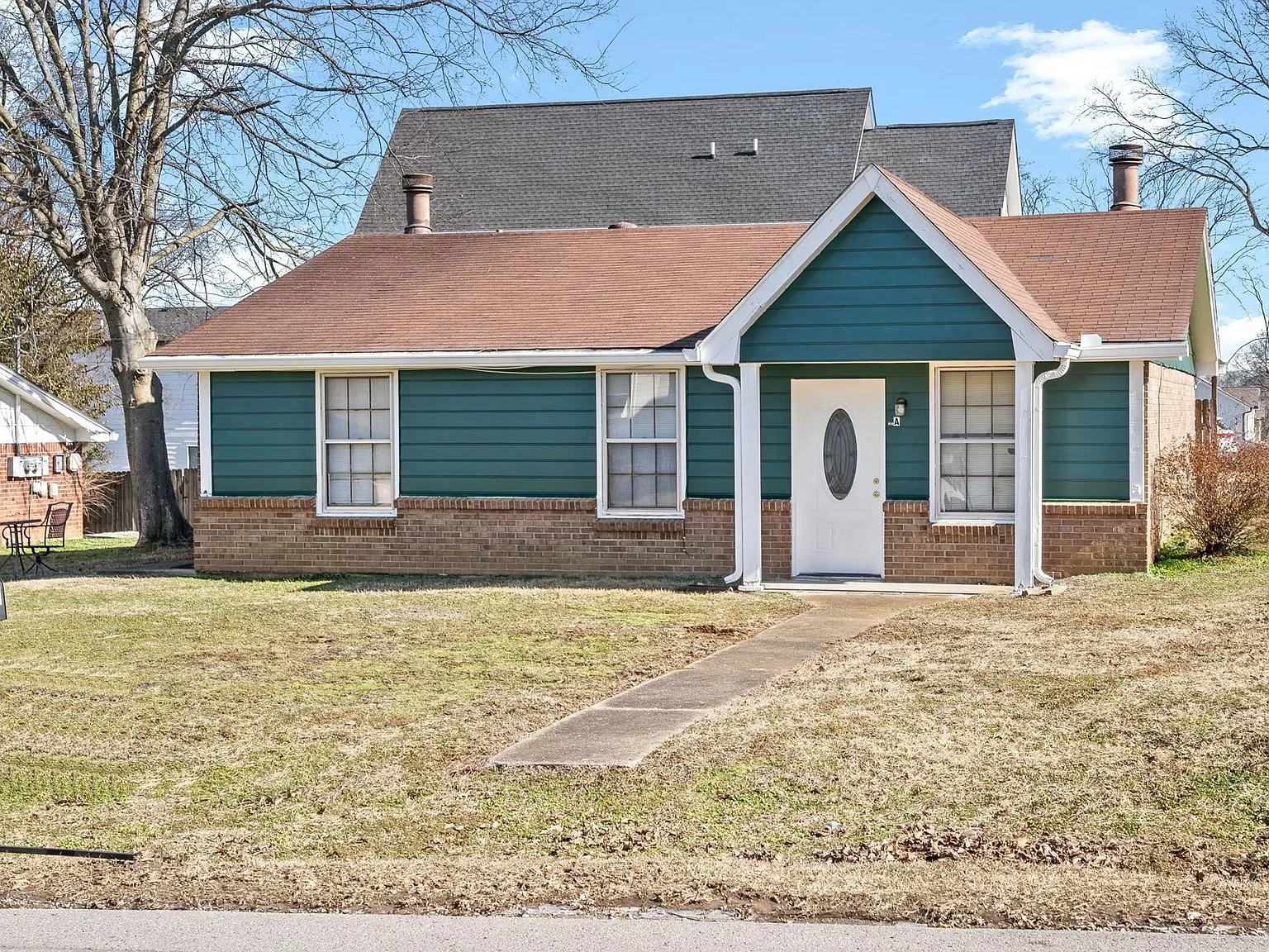 A house with green siding and brick foundation, with a concrete walkway leading to the front door, and a lawn in front.