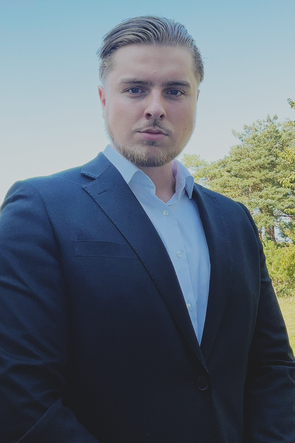 A young man with styled hair, wearing a dark blazer and white shirt, standing outdoors with trees and a clear sky in the background.