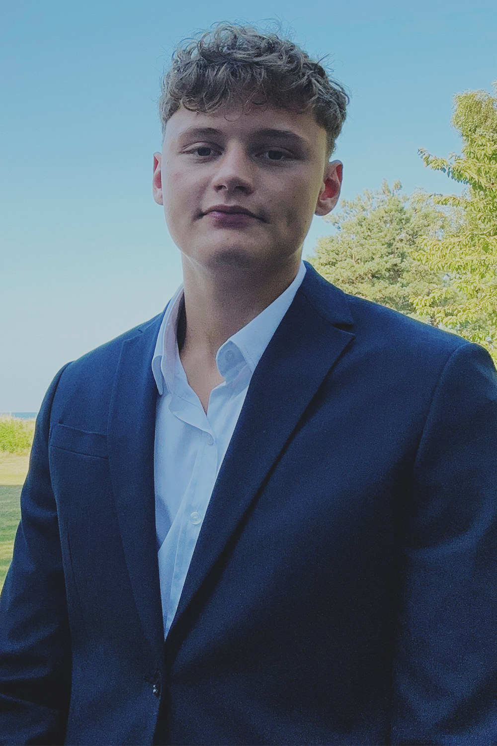 A young man in a blue suit and white shirt standing outdoors near trees with a clear blue sky background.