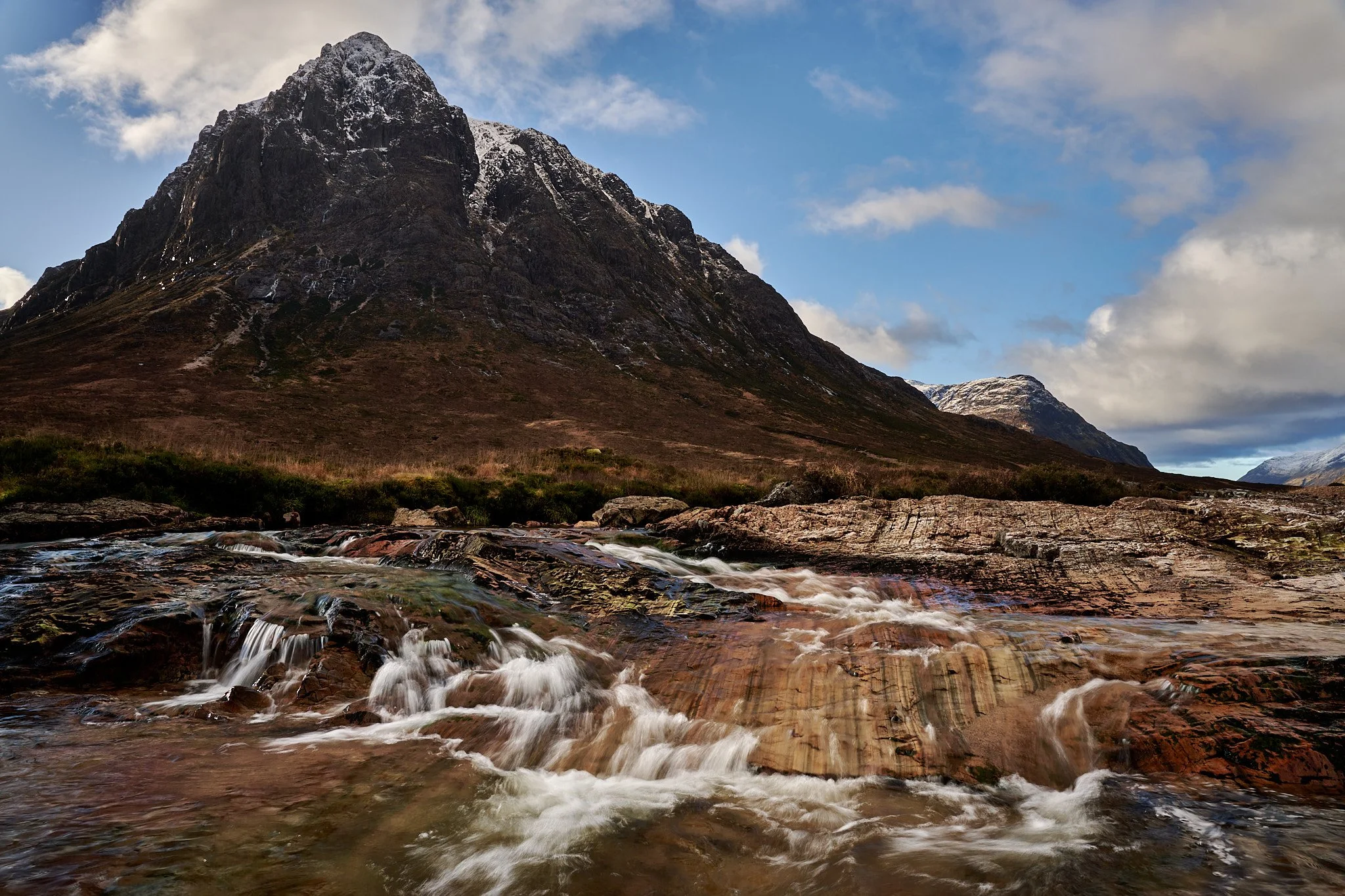 A rugged mountain with a partly snowy peak in the background, a flowing stream in the foreground, and a partly cloudy sky.