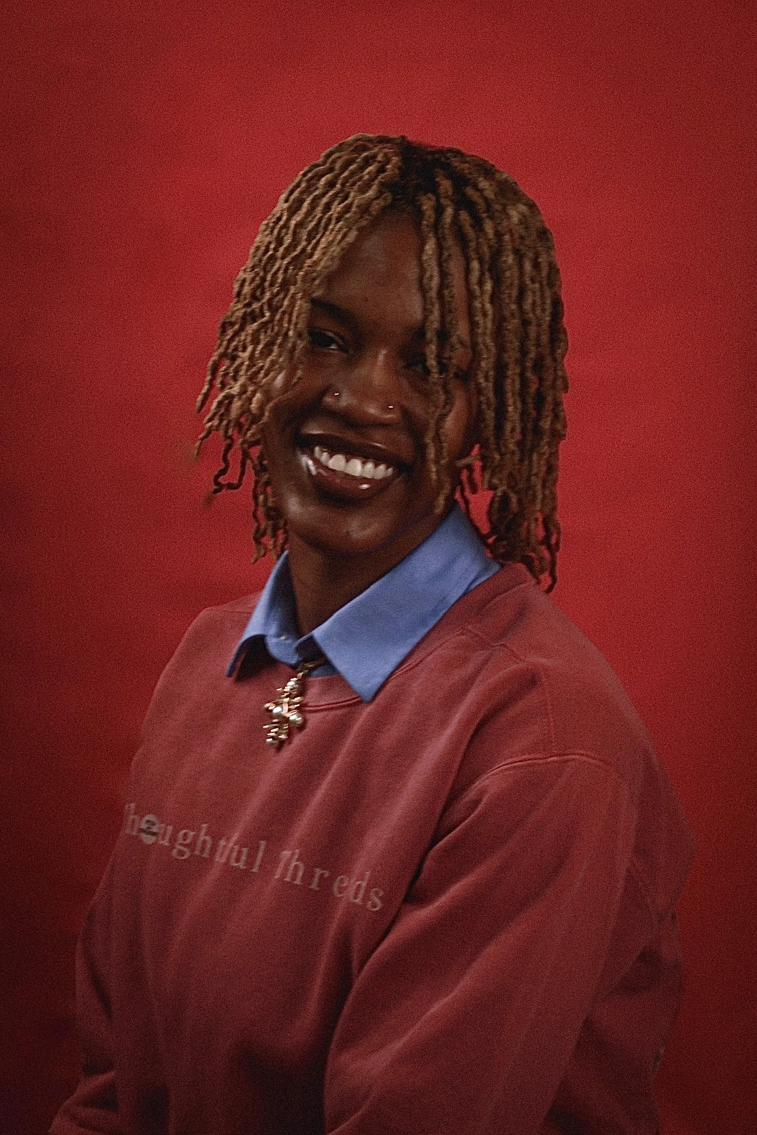A woman with dreadlocks wearing a red sweatshirt with the words "thoughtful threads" and a blue collared shirt underneath, smiling in front of a red background.