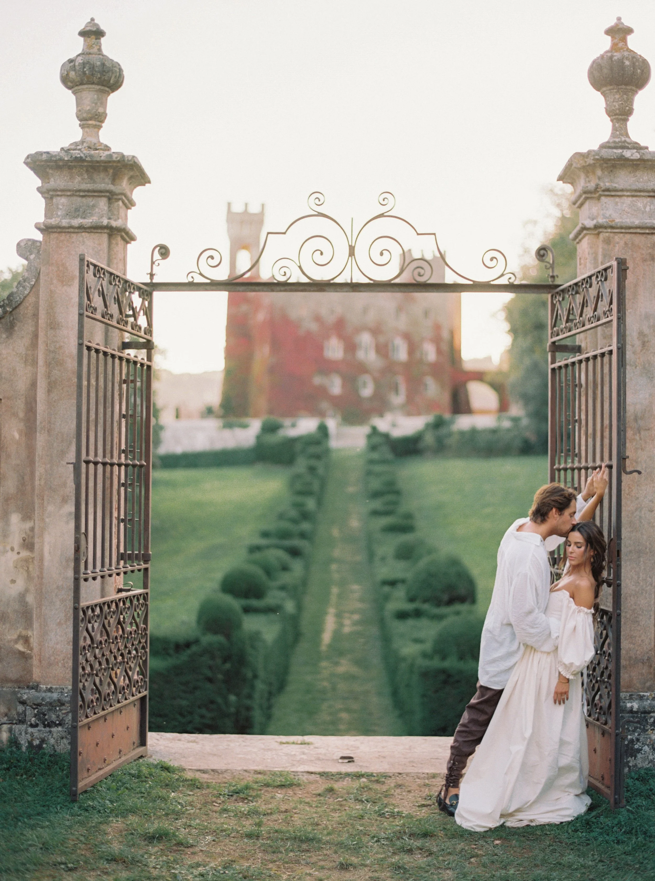 A romantic couple embraced under a wrought iron gate with a castle in the background, surrounded by manicured gardens and greenery.