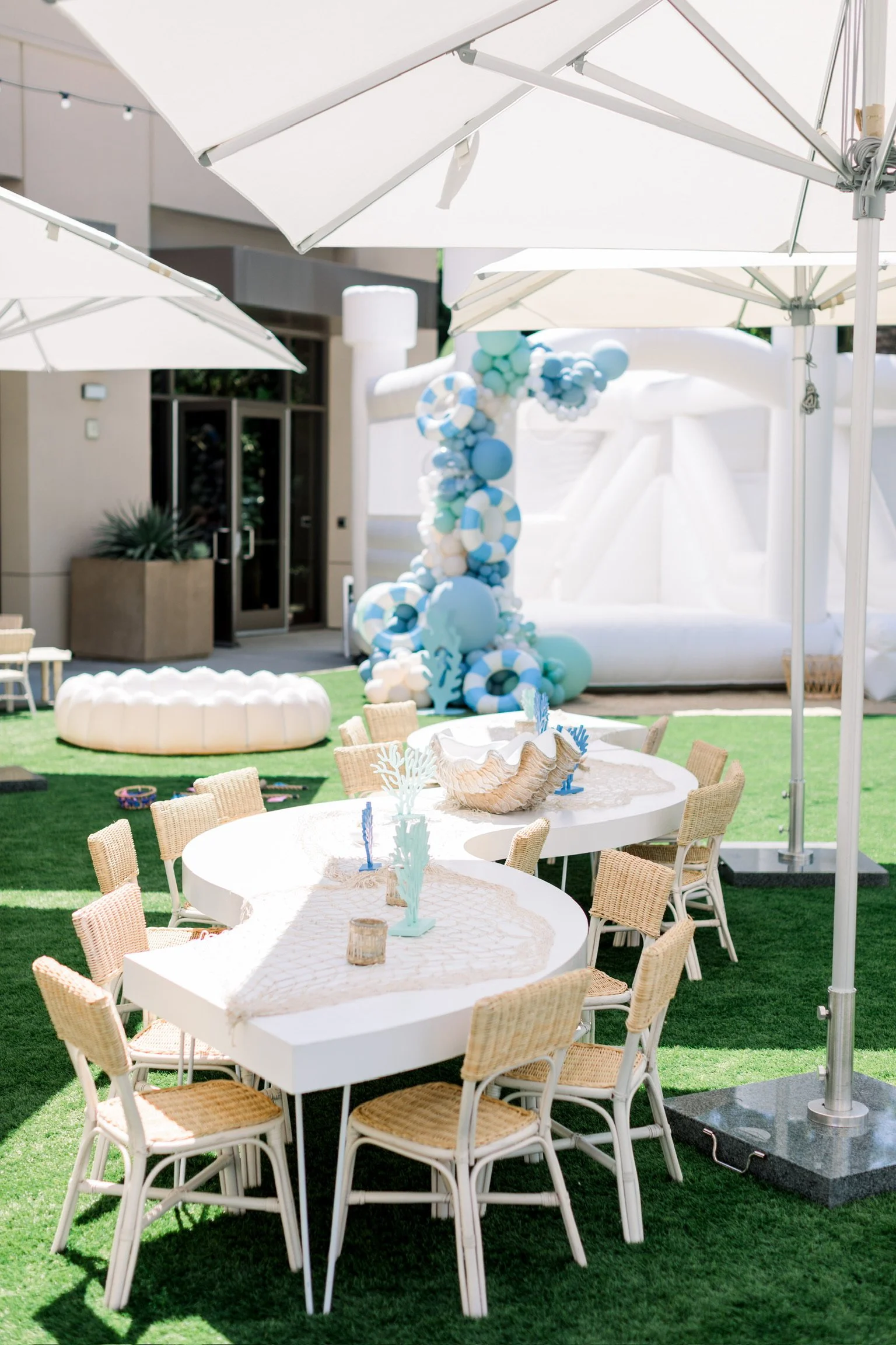 Outdoor party setup with white tables, wicker chairs, umbrellas, a balloon arch with shades of blue and white, and an inflatable water slide in the background.