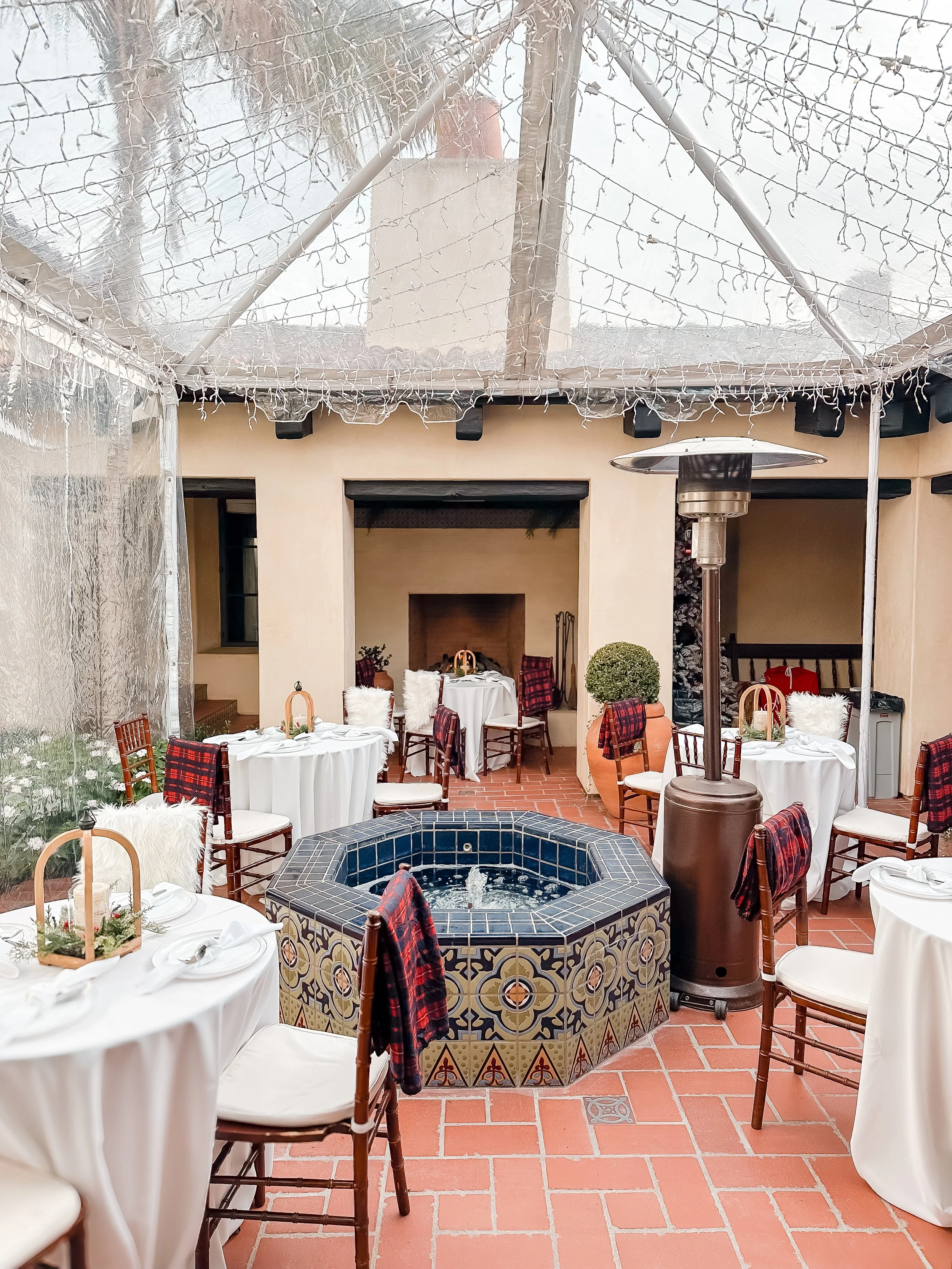 Indoor dining area with round tables covered in white tablecloths, set with plates, silverware, and small centerpieces. Wooden chairs surround the tables, some with plaid scarves draped over the backs. In the center of the room is a tiled hot tub or fountain. The space is decorated with potted plants and features a brick fireplace in the background. A clear plastic tent with string lights is overhead, and patio heaters are present.