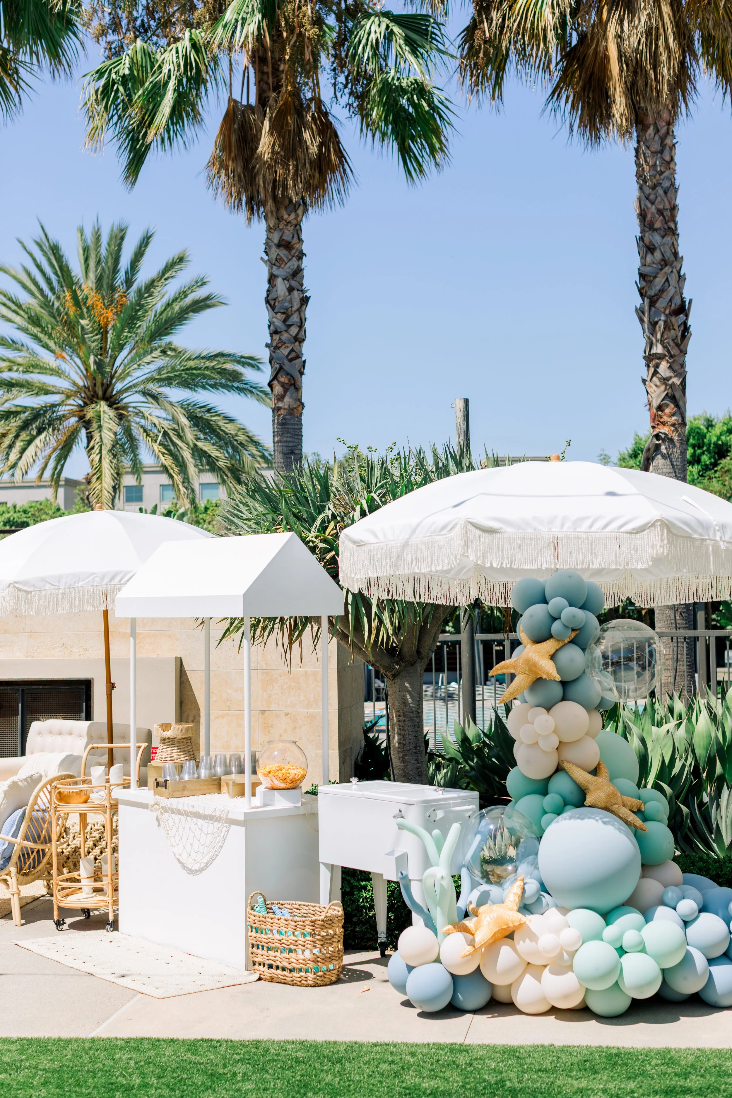 Decorative outdoor setup with white umbrellas, pastel balloons, and tropical palm trees.