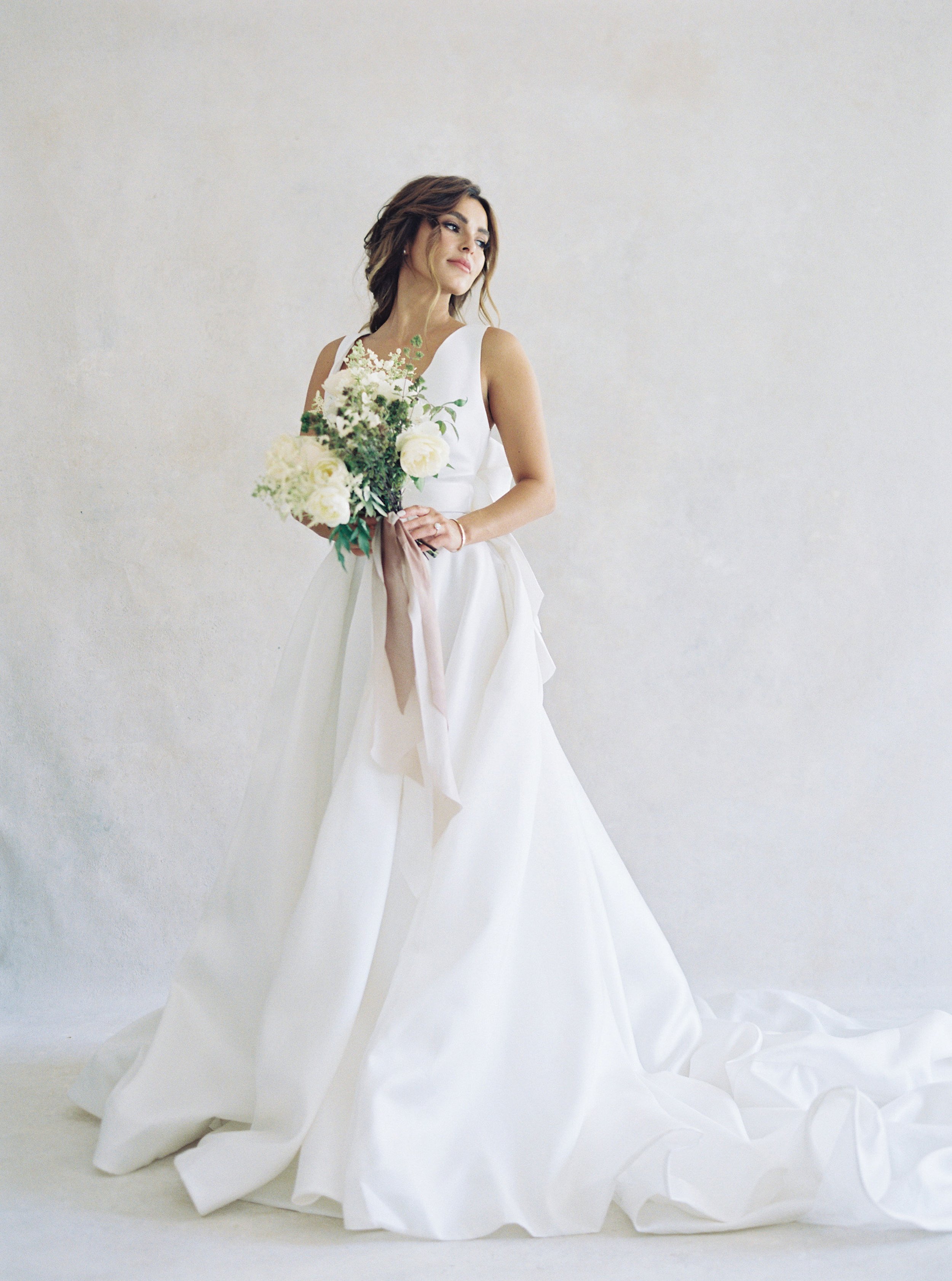 A woman in a white wedding gown holding a bouquet of flowers against a plain background.