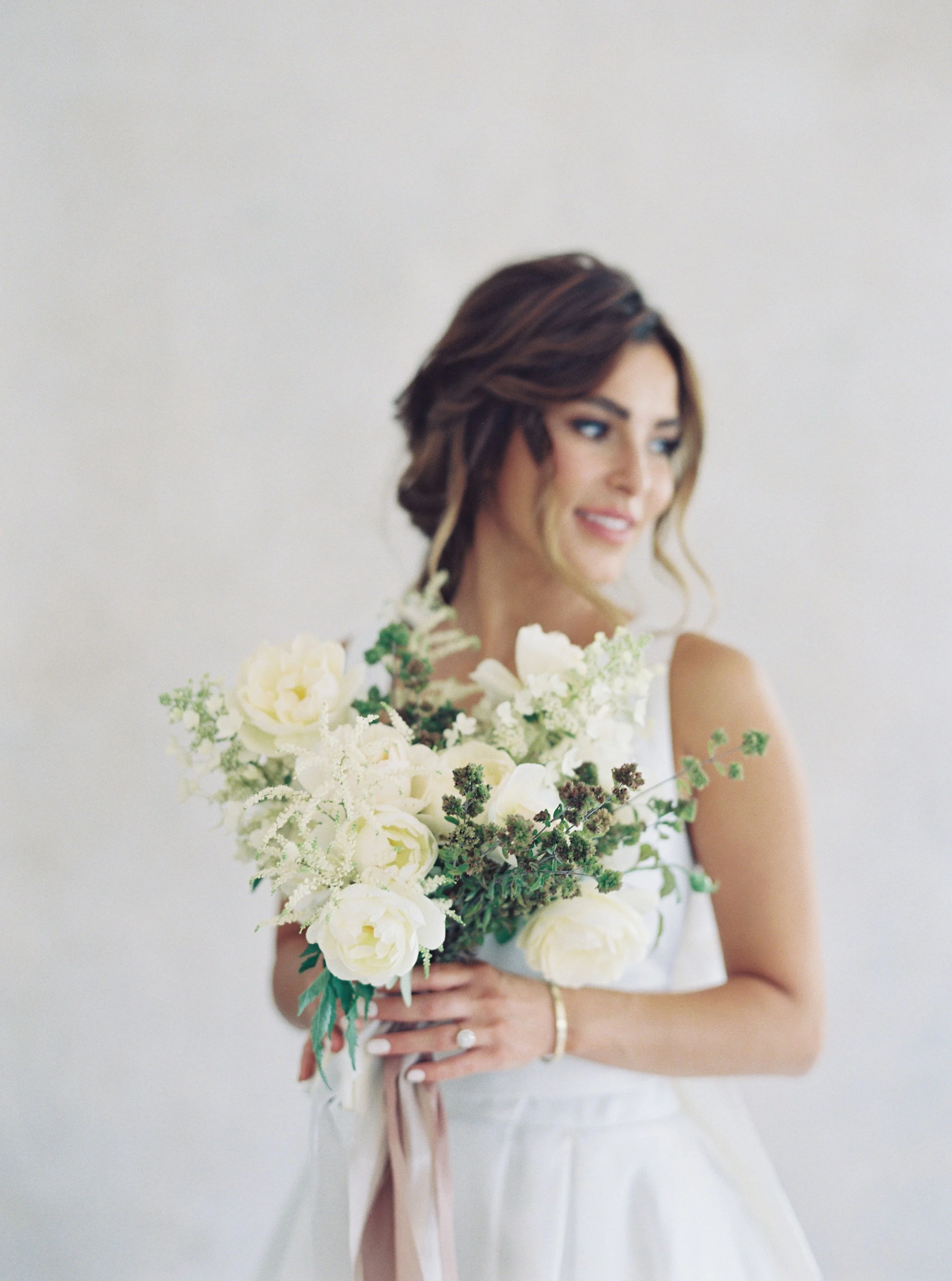 A woman in a white dress holding a bouquet of white flowers, with brown hair styled in loose curls, smiling softly against a plain light background.