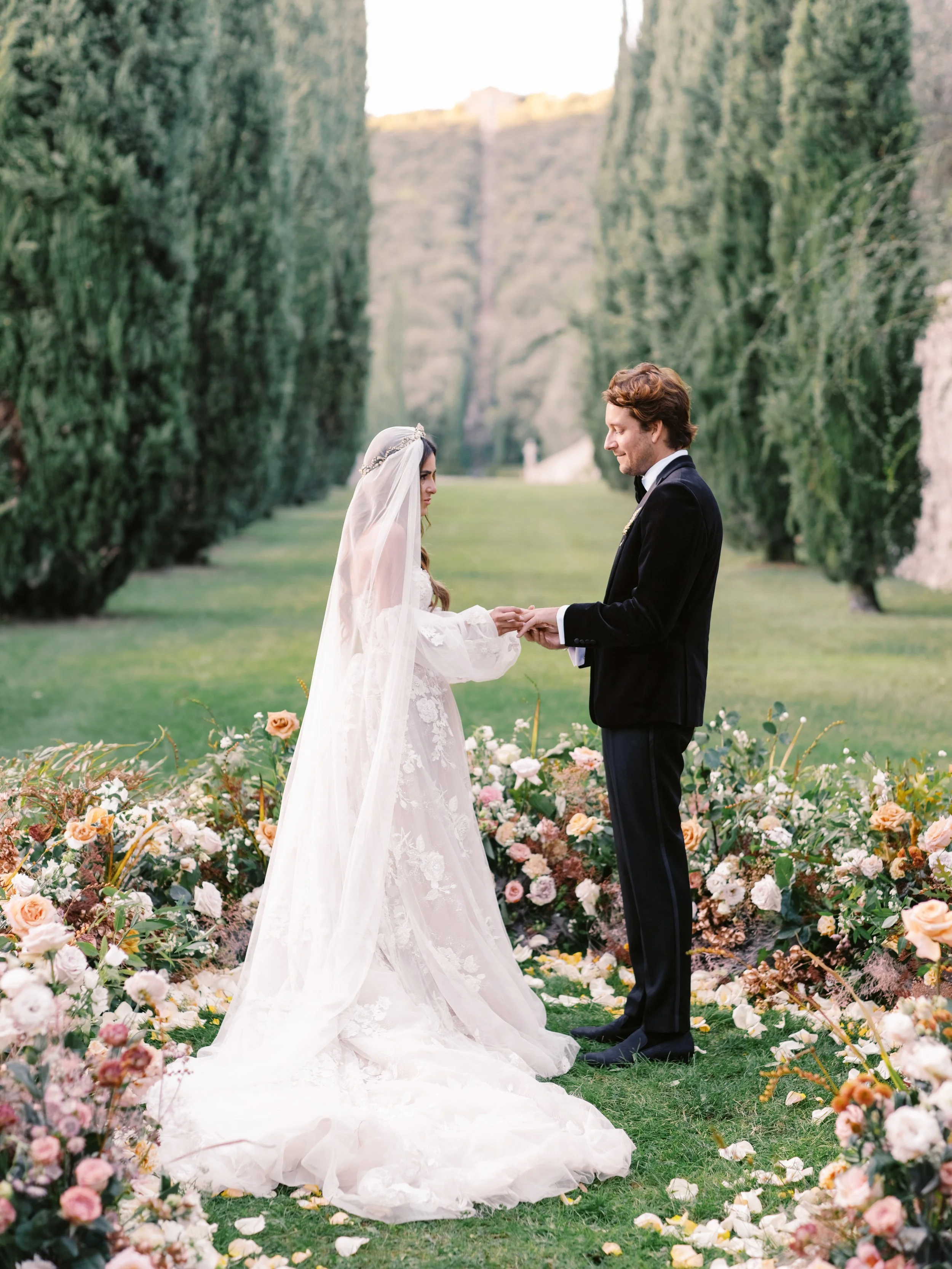 A bride and groom holding hands in a garden with flowers, trees, and green grass.
