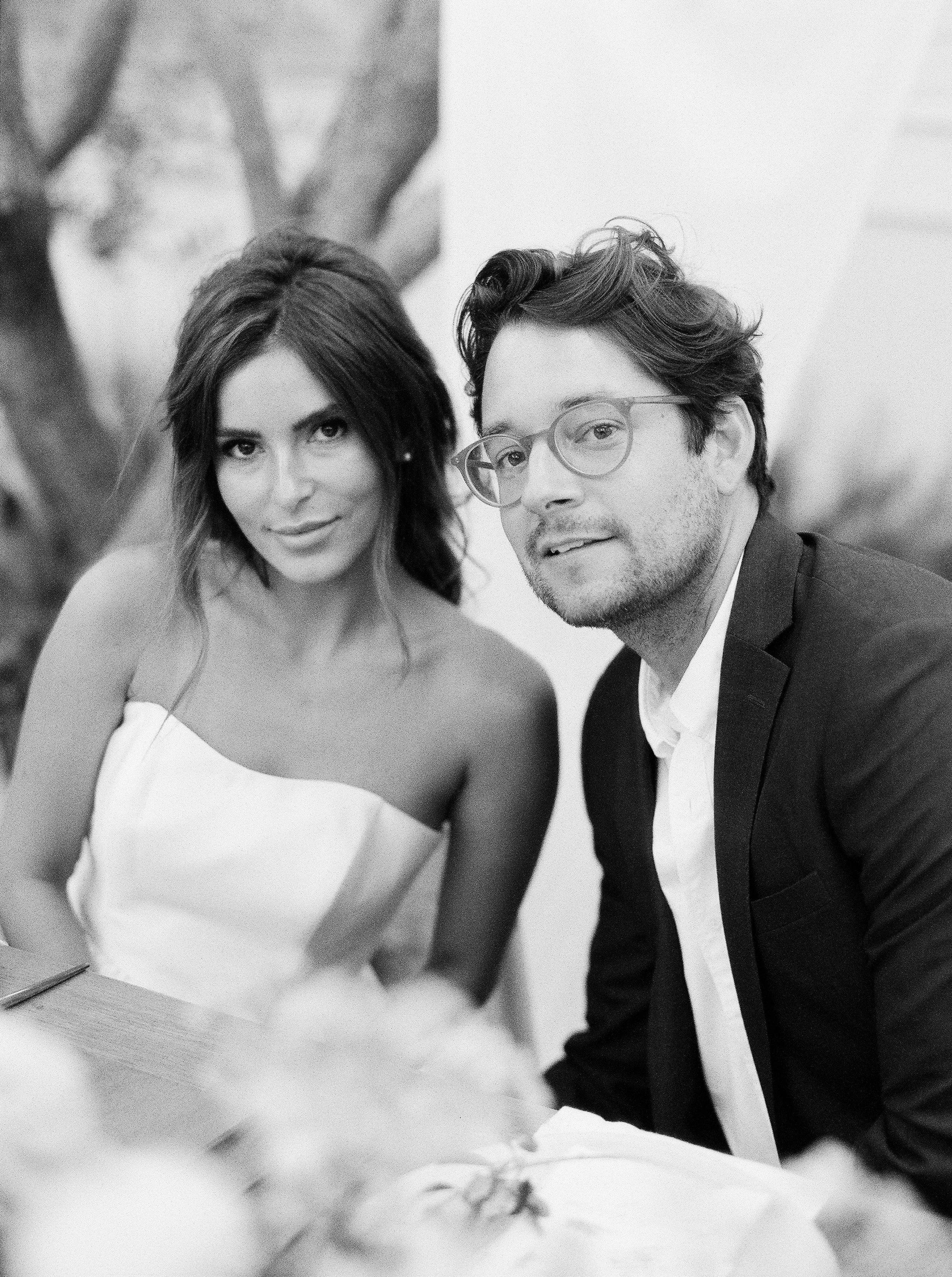 A black and white photo of a man and a woman sitting at a table, with the woman wearing a strapless dress and the man in a suit with glasses. They are looking at the camera.