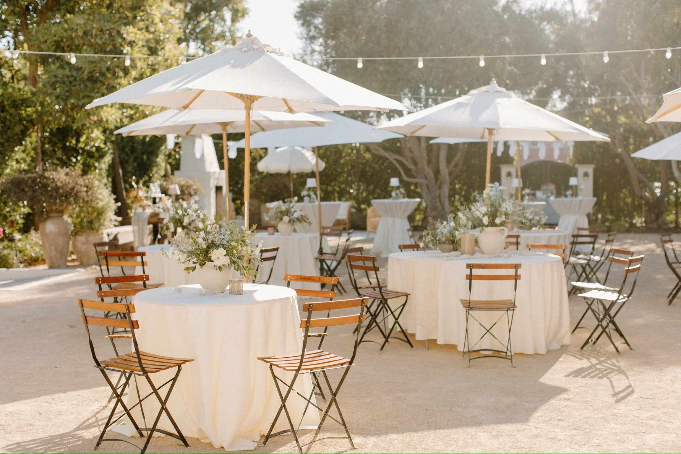 Outdoor wedding reception setup with round tables covered in white tablecloths, decorated with floral centerpieces and surrounded by wooden chairs. Large white umbrellas provide shade, and string lights are hung overhead in a lush garden setting.