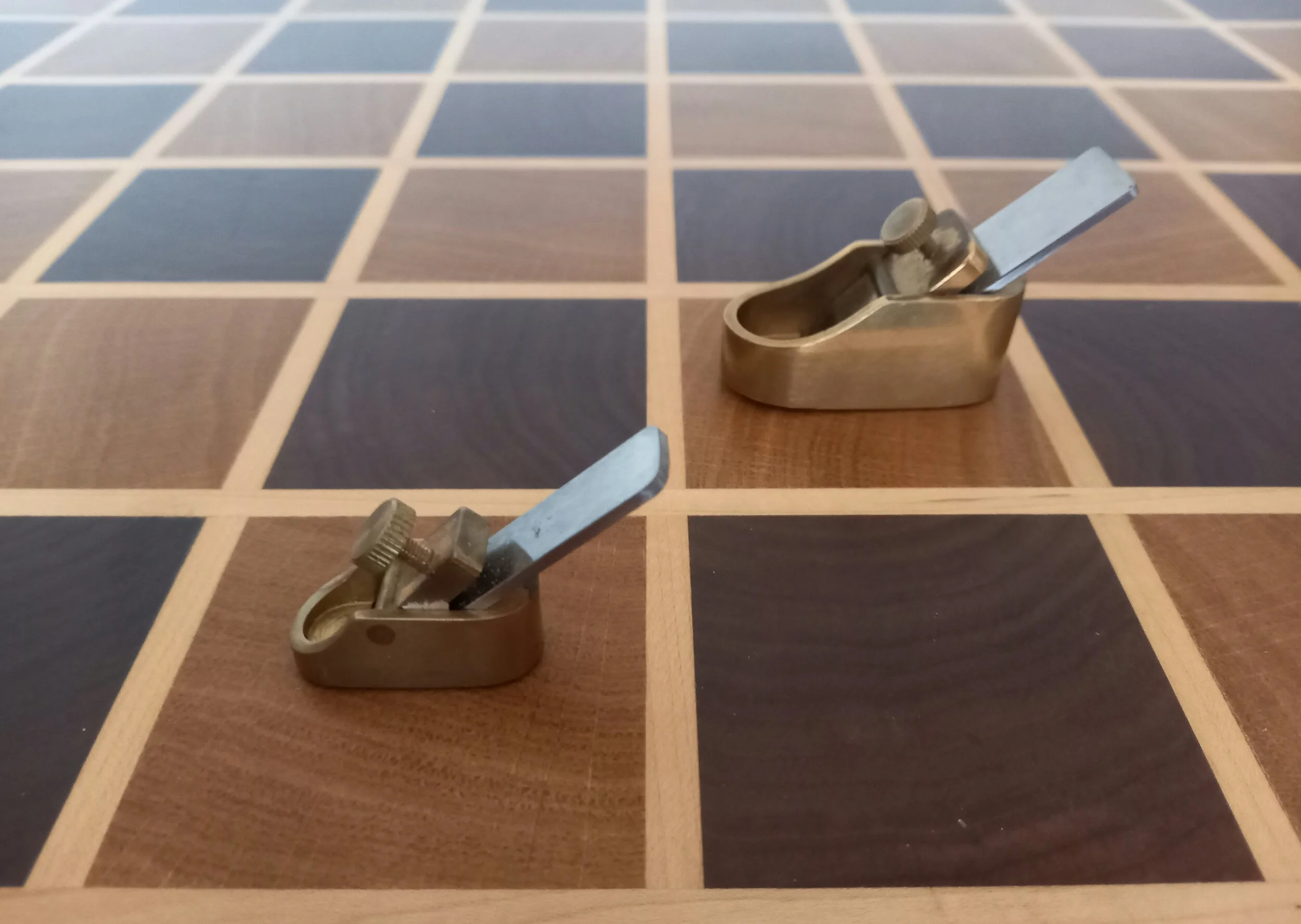 Close-up of two brass thumb planes on a wooden chessboard with black and brown squares.