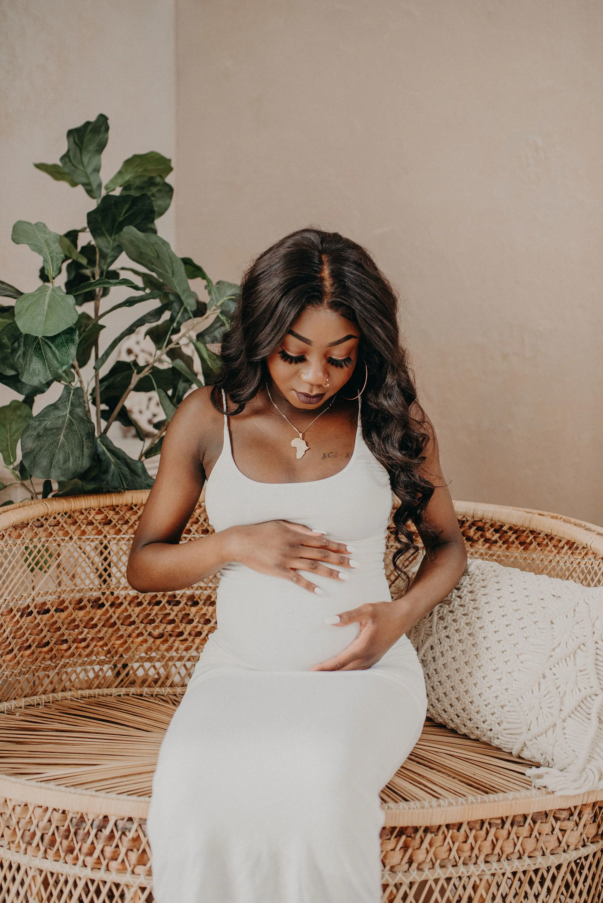 Pregnant woman in a white dress resting on a wicker daybed beside a leafy houseplant