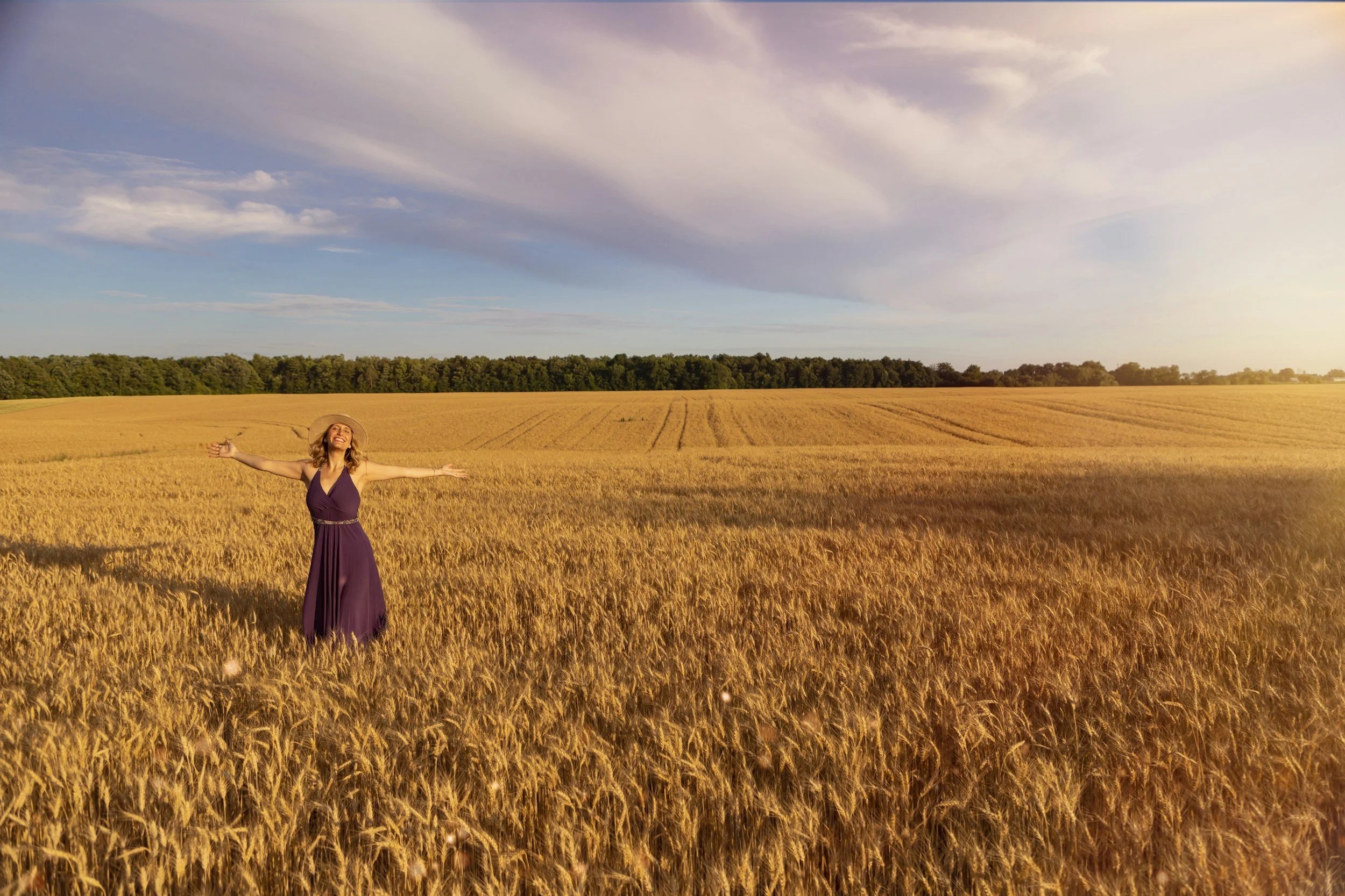 Woman in a purple dress and sun hat standing in a rural landscape with arms outstretched in a golden wheat field at sunset
