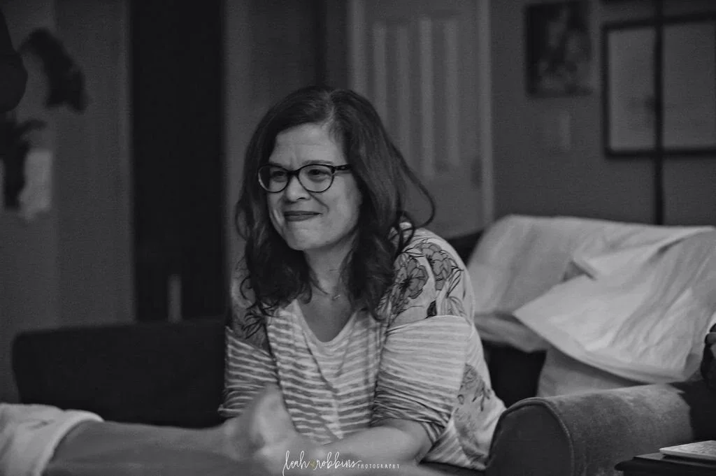 Smiling woman wearing glasses and a striped shirt seated on a couch in a black-and-white portrait