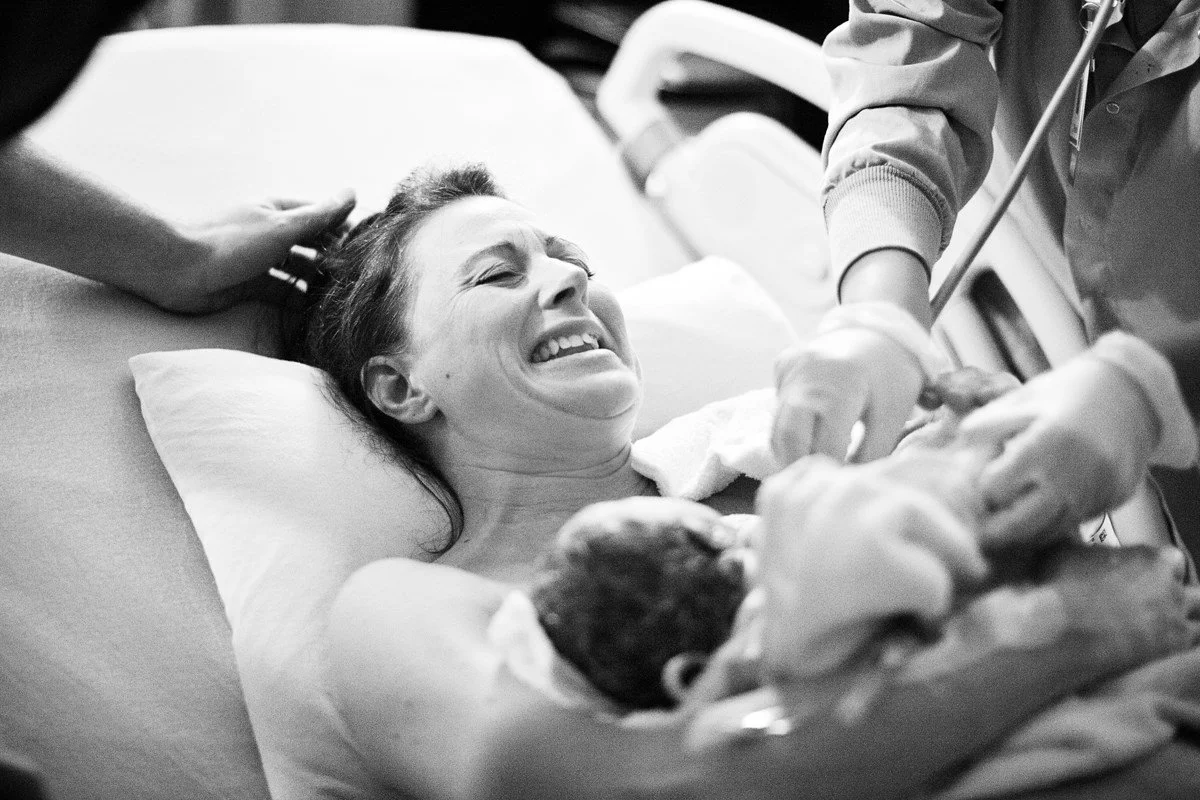 Smiling mother holding her newborn baby in a hospital bed as a healthcare provider assists.