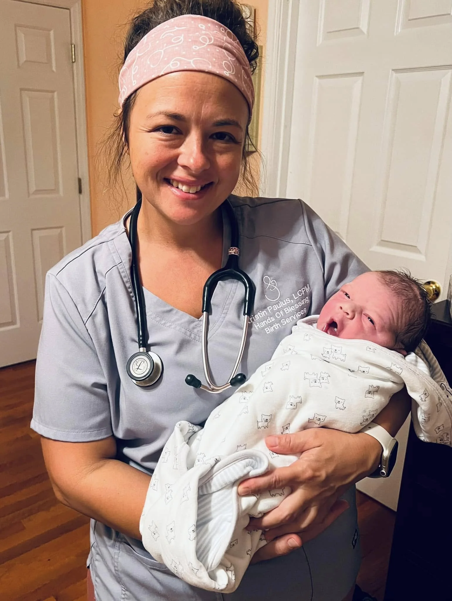 Smiling midwife in gray scrubs and a pink headband holds a newborn baby wrapped in a patterned blanket.