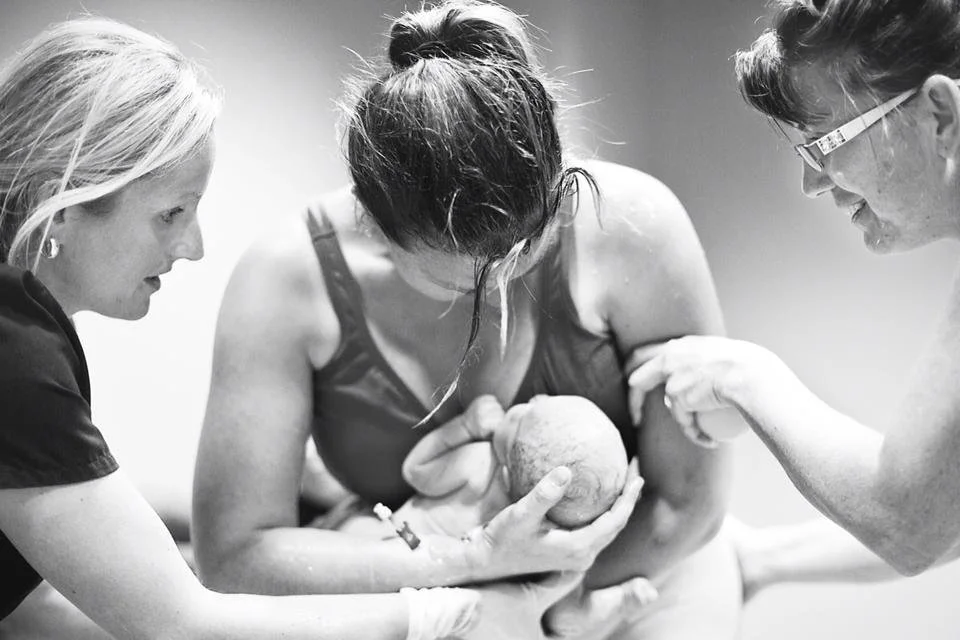 Black-and-white photo of a mother holding her newborn baby during a supported water birth, assisted by two caregivers.