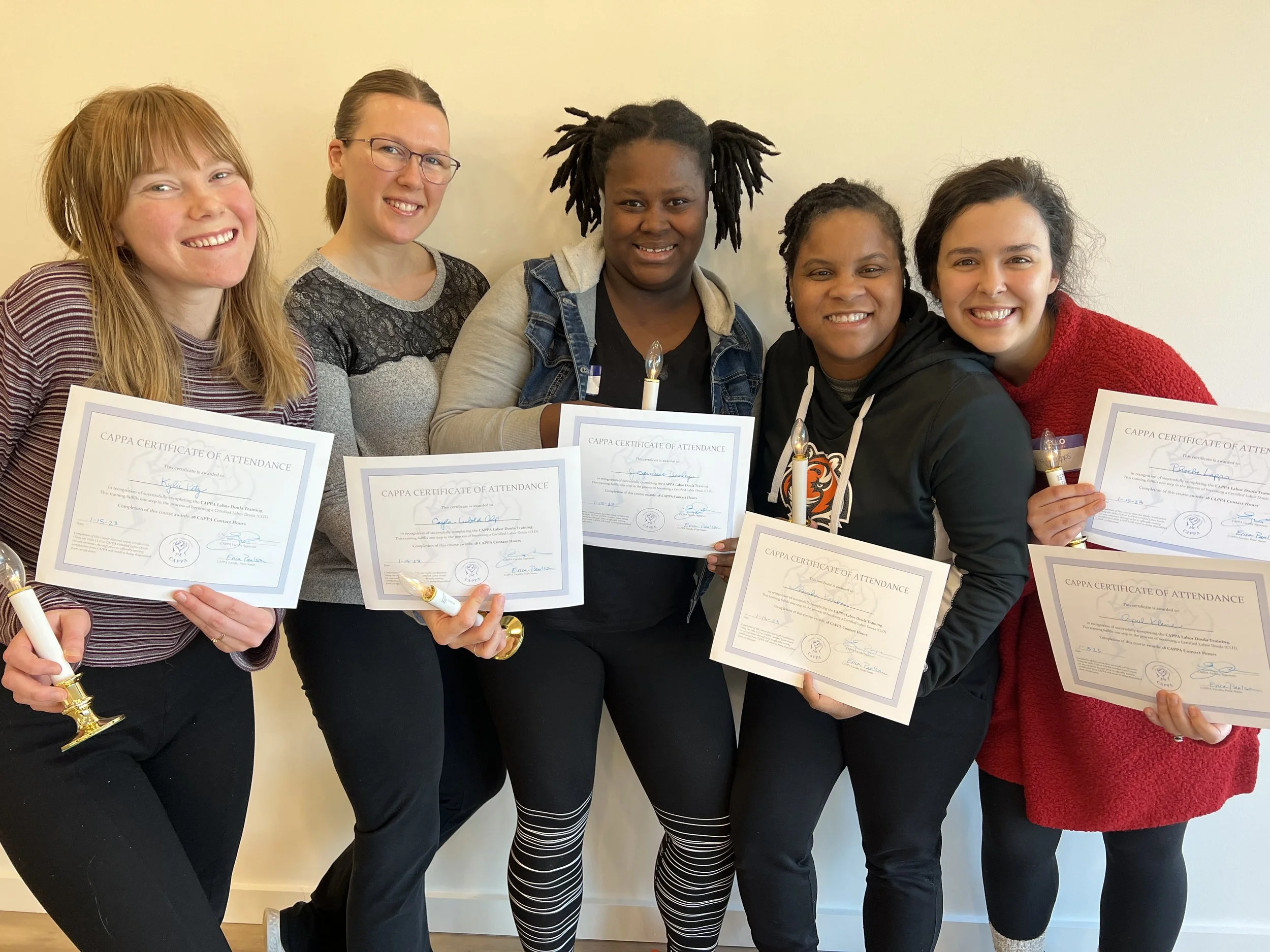 Five smiling women hold doula certificates of attendance and celebratory candles after completing labor doula training.