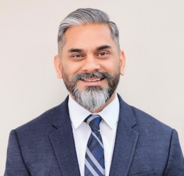 Vik Singh, dressed in a navy blazer, white shirt, and striped tie against a plain light background.
