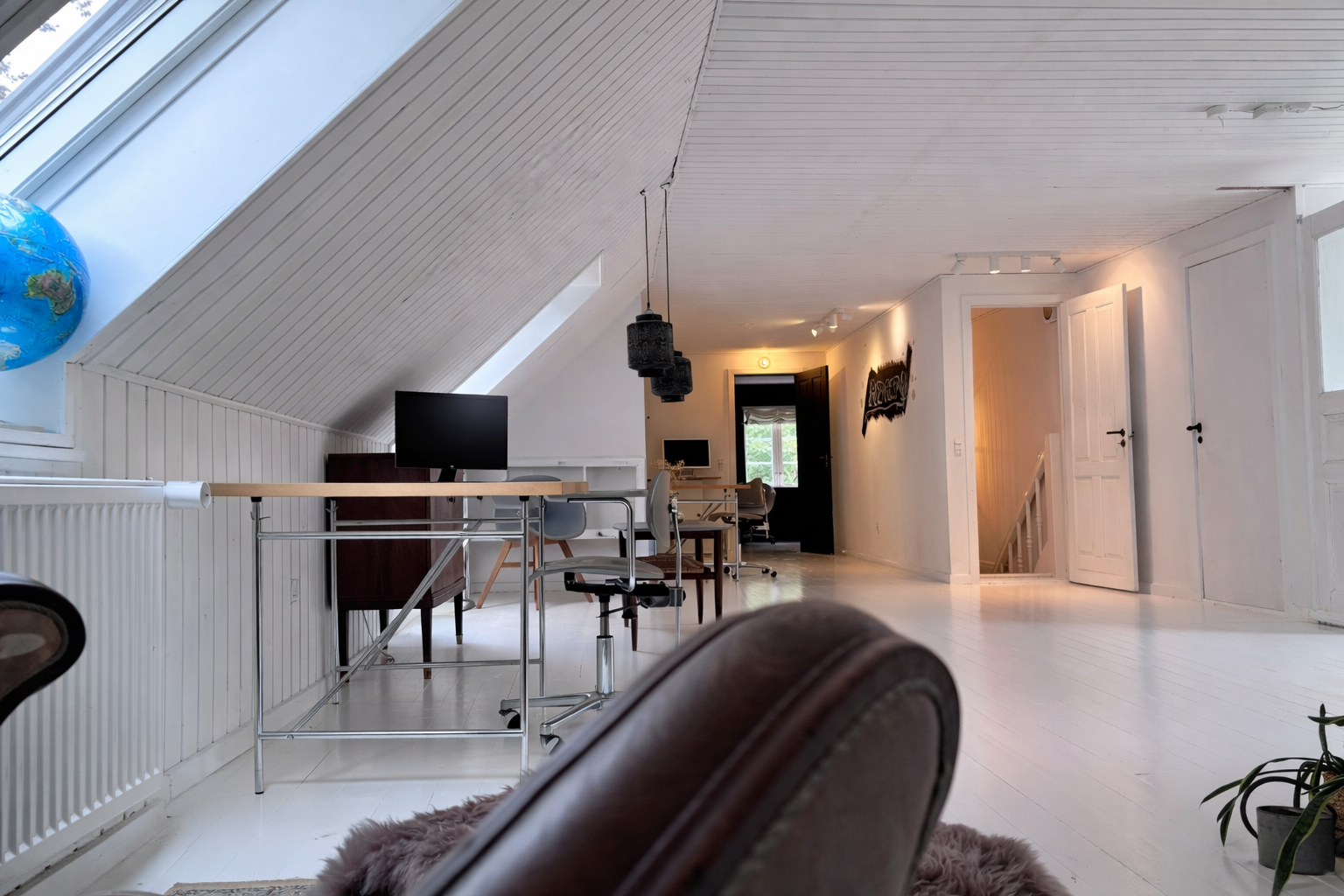 Heller Working-Loft-Bereich unter schrägem Dach mit weißem Holzboden, Egon Eiermann Schreibtisch mit Monitor, Drehstuhl, Oberlichtfenster und weiterem Arbeitsplatz im Hintergrund.