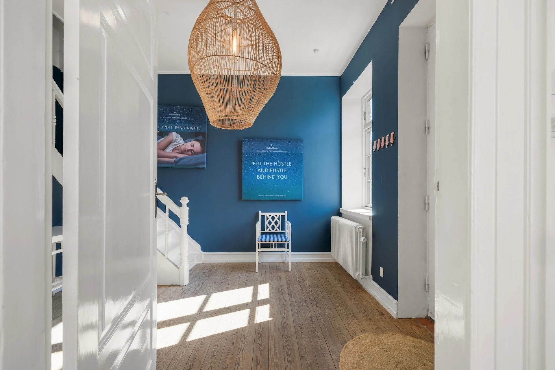 großzügiges Treppenfoyer des Alten Pfarrhaus auf Ærø. mit hellem Boden, blauen Wänden und weißen Türen und weißer Treppe. Ein Stuhl steht im Raum, an der Decke hängt eine Lampe in Form einer Reuse.