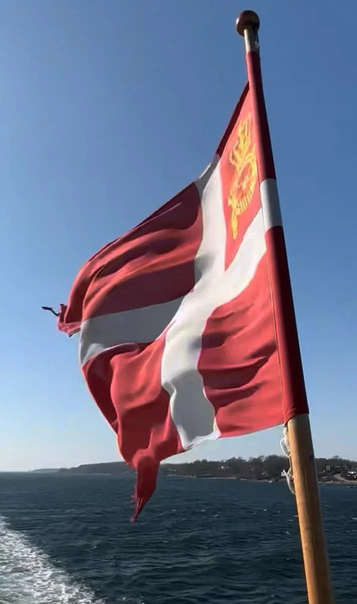 Dänische Flagge, Rot mit weißem Kreuz und einem Wappen im oberen Eck, weht an einer Holzstange vor blauem Himmel im Hafen von Ærø.