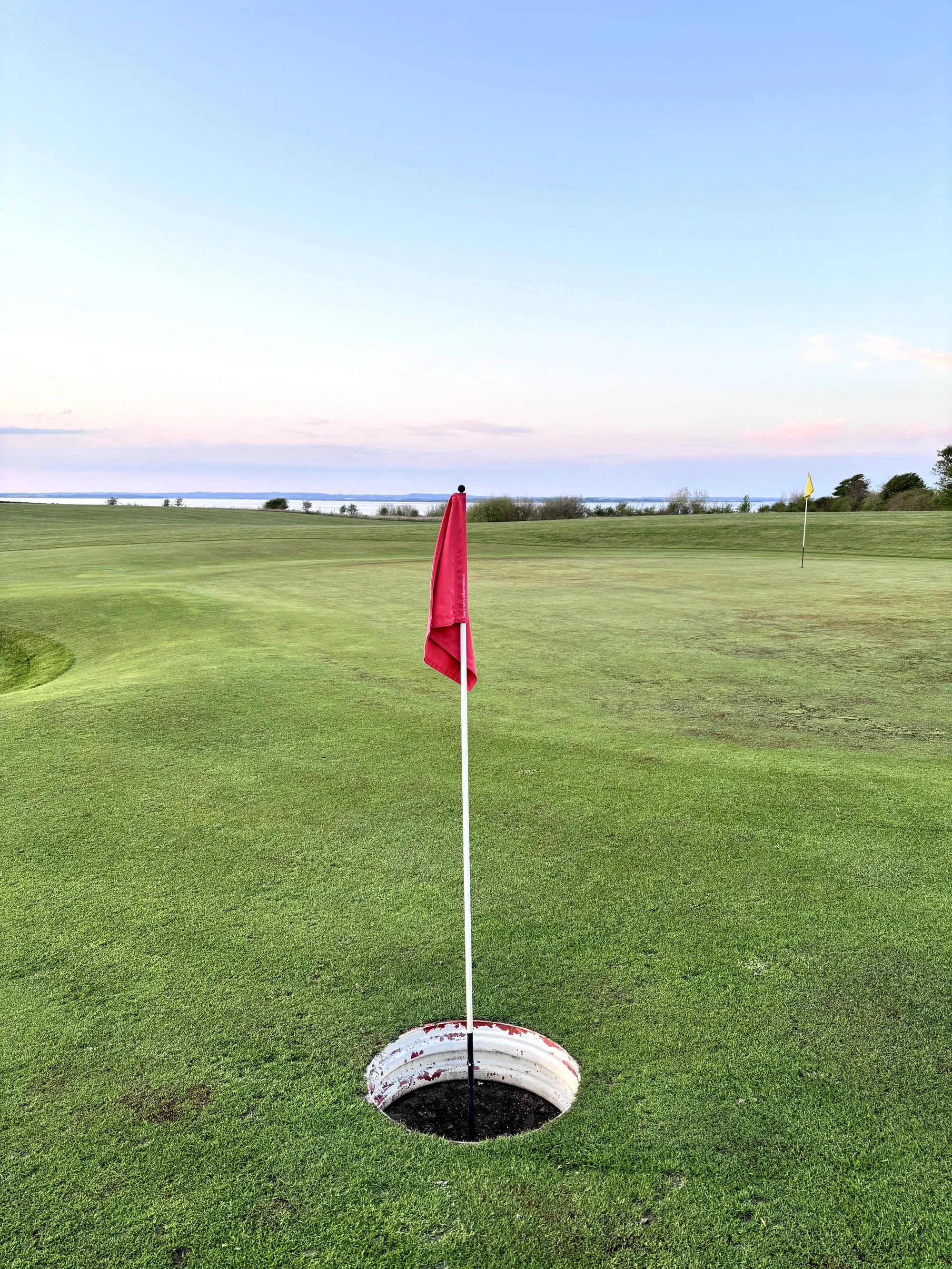 Golfplatz mit einem großen Foot-Golf-Loch, indem eine rote Lochfahne steckt. Am Horizont das ruhige Meer und rötlich blauer Himmel
