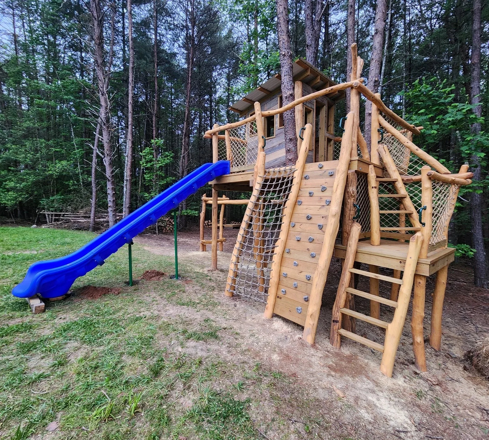 Custom treehouse playground with an 8-foot-tall play deck, slide, climbing wall, cargo net, double ladder, monkey bars, swing set, and zipline at Cottage Park in Morganton, North Carolina.