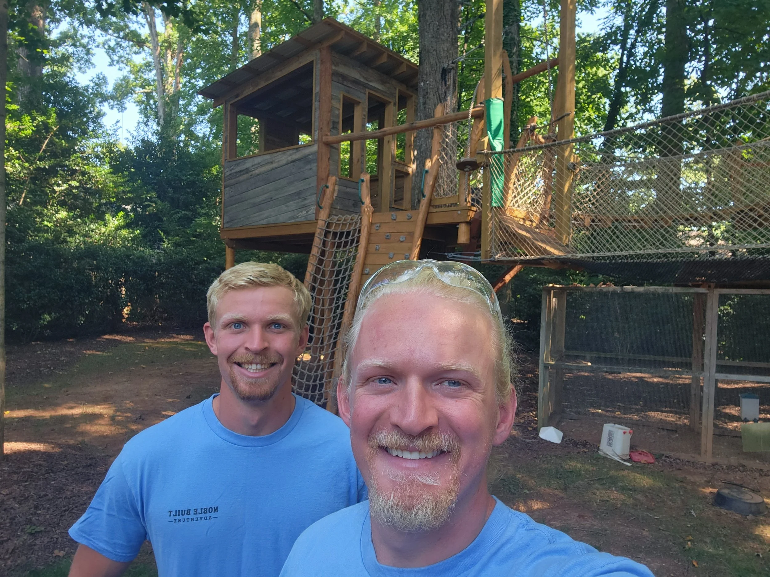Two men smiling outdoors on a sunny day, with a treehouse structure and trees in the background.