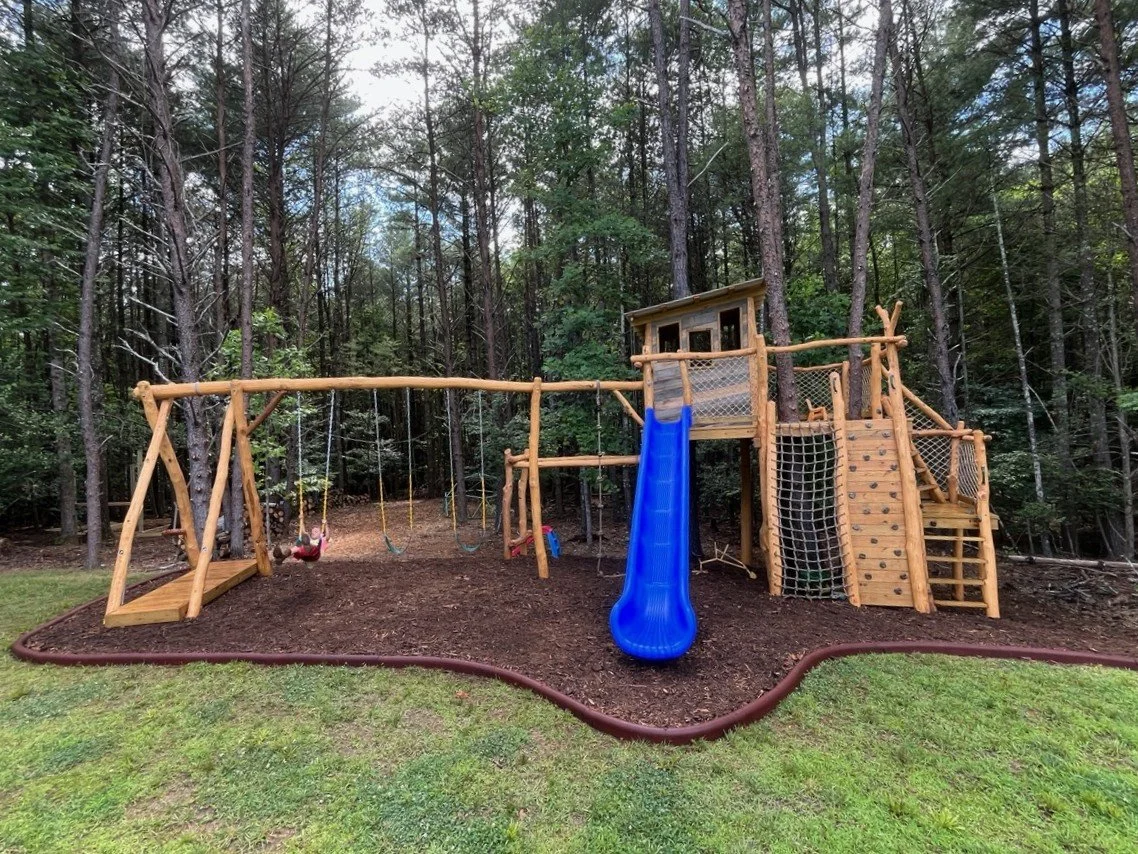  Custom treehouse playground with an 8-foot-tall play deck, slide, climbing wall, cargo net, double ladder, monkey bars, swing set, zipline, and mulched play area at Cottage Park in Morganton, North Carolina.
