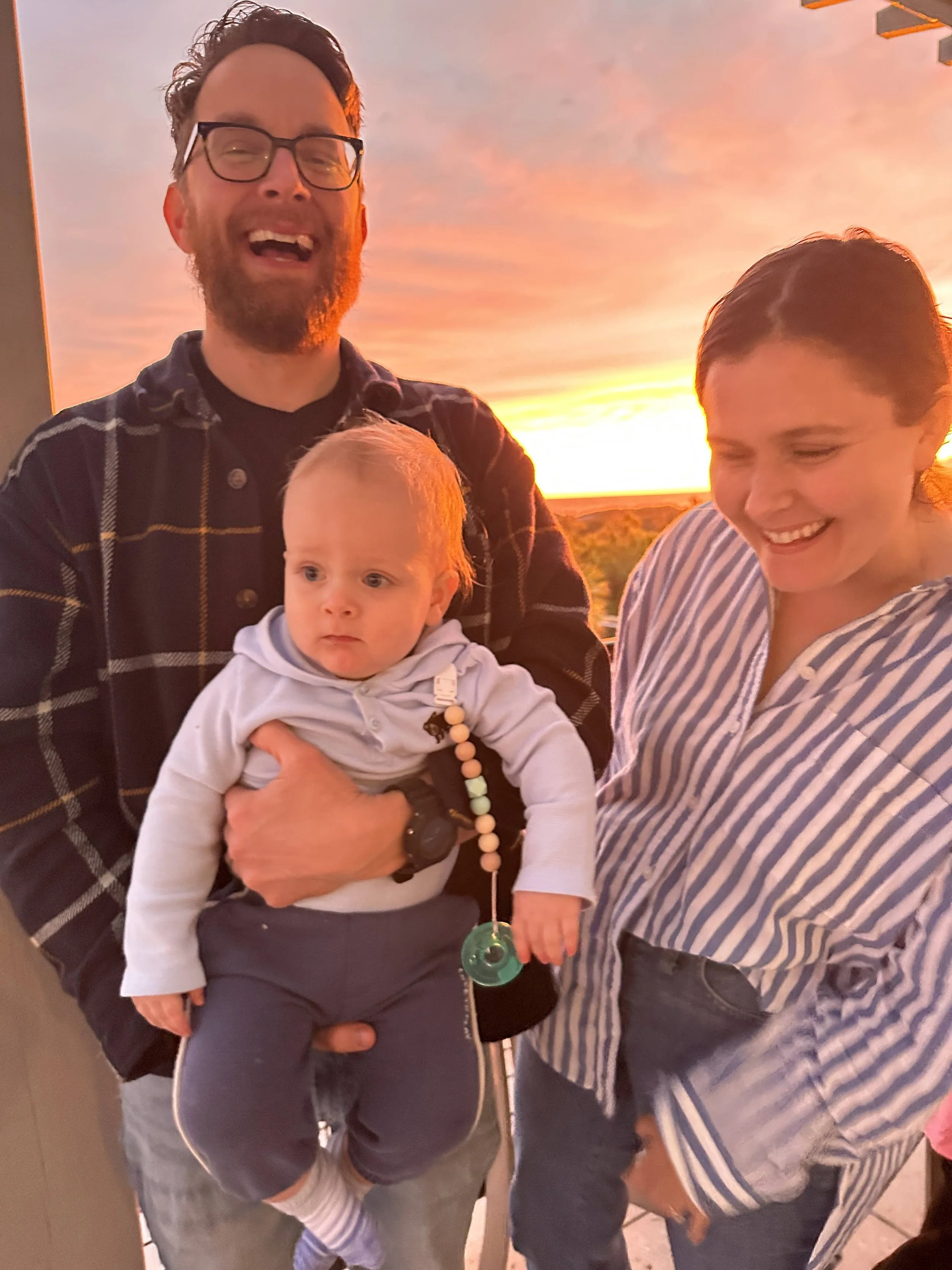 A happy man with glasses holding a baby, standing next to a smiling woman, against a sunset background.