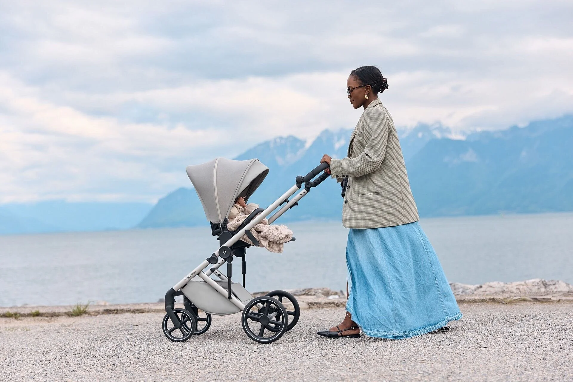 Une femme portant un blazer gris et une longue jupe bleue pousse une poussette sur un sentier au bord du lac. Capturée à Montreux par un photographe professionnel.