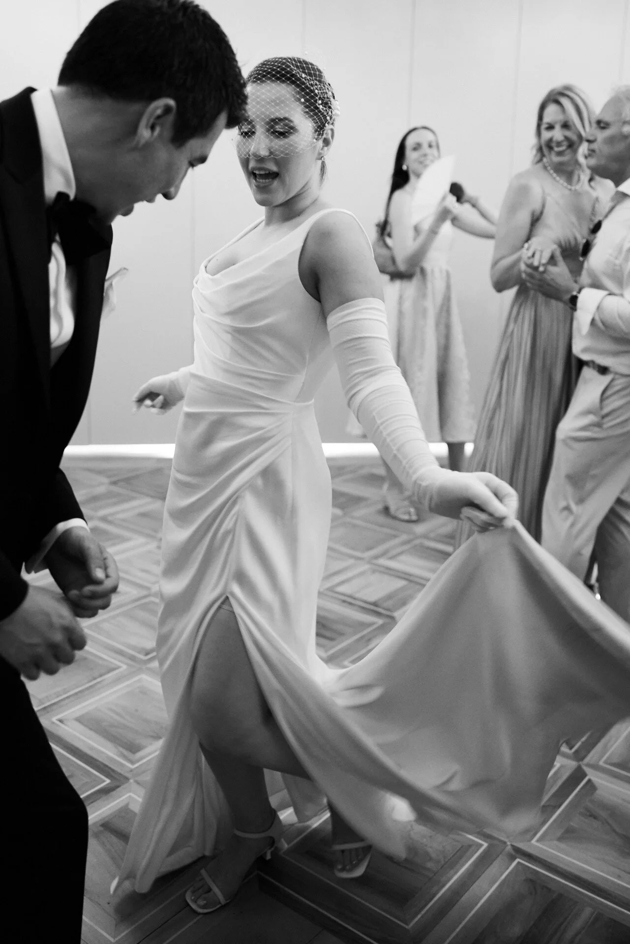 Une femme en robe de mariée qui danse avec un homme lors d'une réception, entourée d'invités qui rient et les regardent.