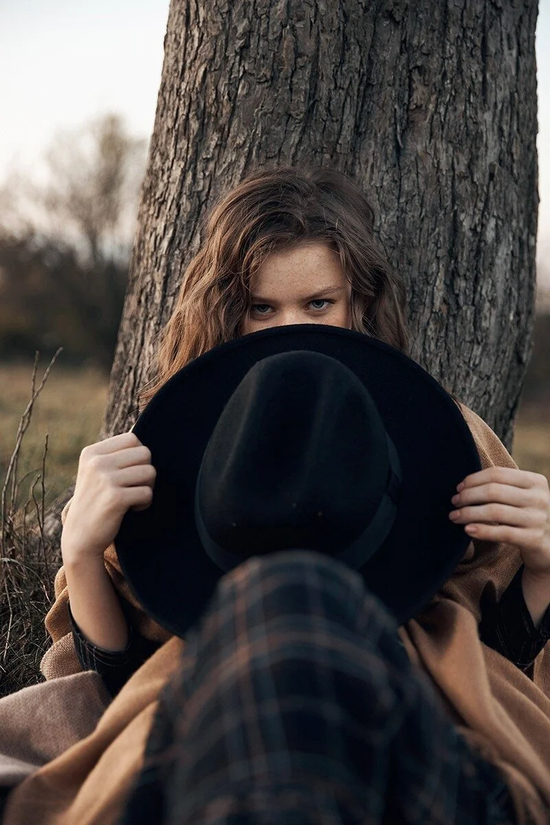 Young_woman_with_black_hat_sitting_by_a_tree.jpg