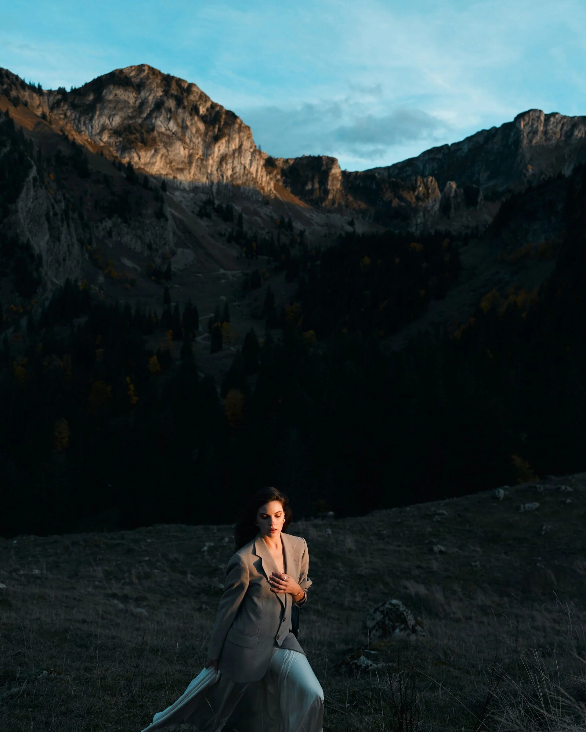 Une femme portant un tailleur gris et une jupe blanche pose dans un paysage montagneux au coucher du soleil. Capturée à Montreux par un photographe professionnel.