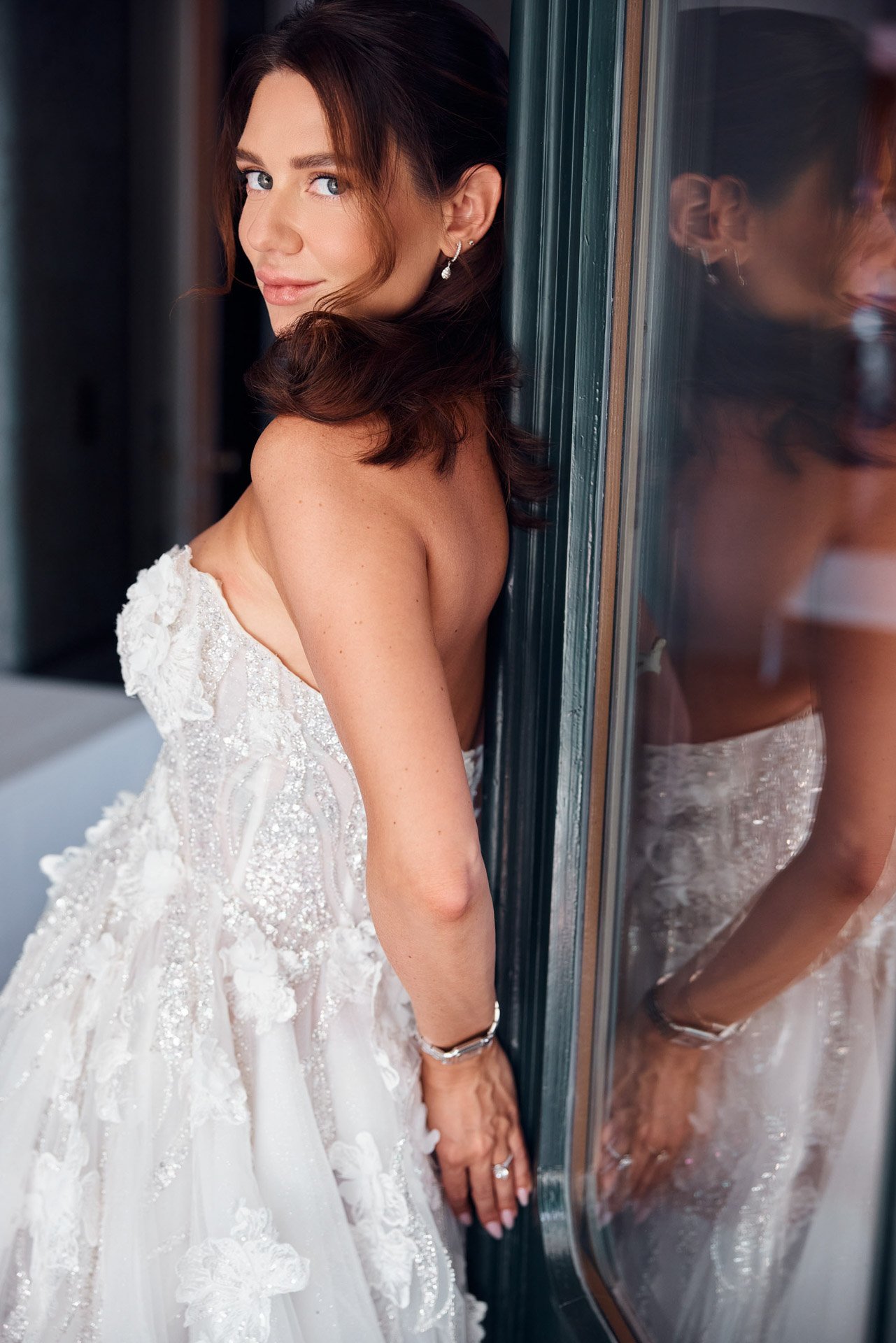 Une mariée en robe blanche étincelante, appuyée contre une fenêtre.jpeg
