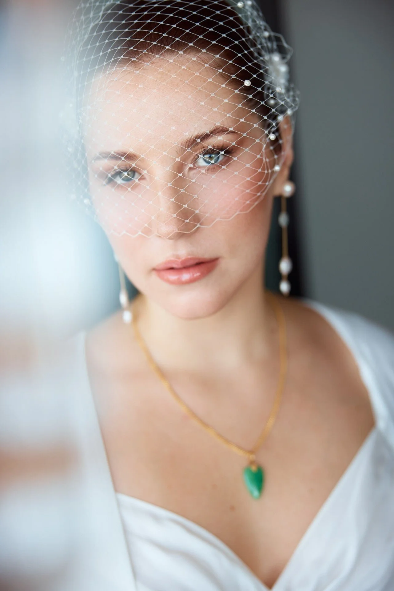 Une femme aux yeux bleus, maquillée, portant un voile « cage à oiseaux » orné de perles, des boucles d'oreilles, un collier en or et un pendentif vert, vêtue d'un haut blanc.