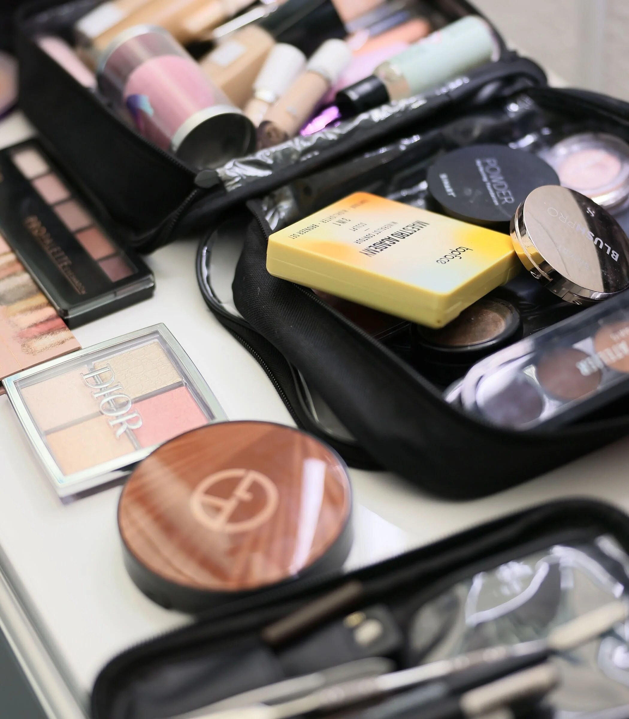 Open makeup bag with various cosmetics, including blush, setting spray, and foundation bottles, on a white surface.