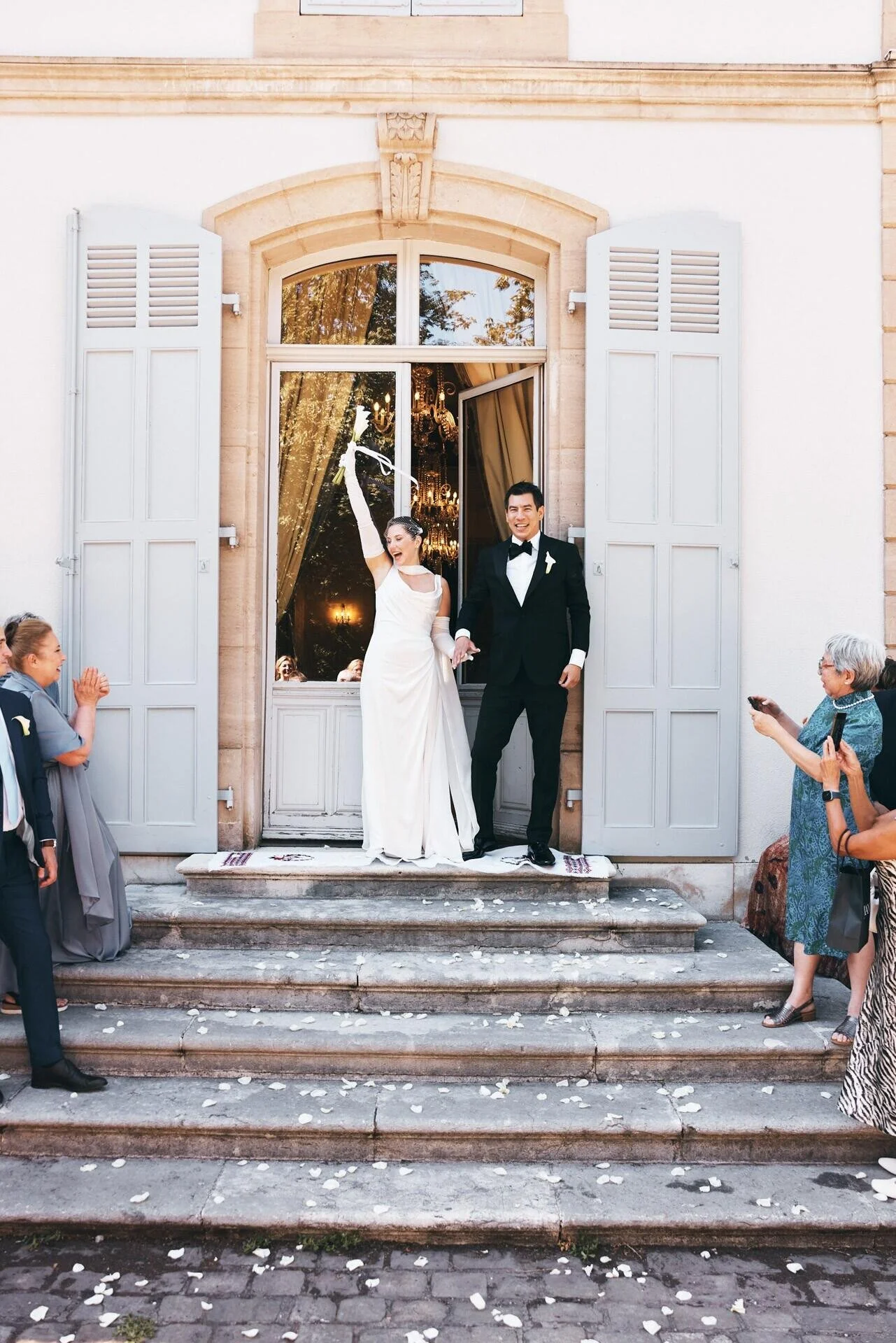 Un couple de jeunes mariés sort d'un immeuble, tout sourire, main dans la main. La mariée porte une robe blanche et un gant, tandis que le marié est vêtu d'un smoking noir. Les invités font la fête sur les marches en lançant des pétales de fleurs blanches.