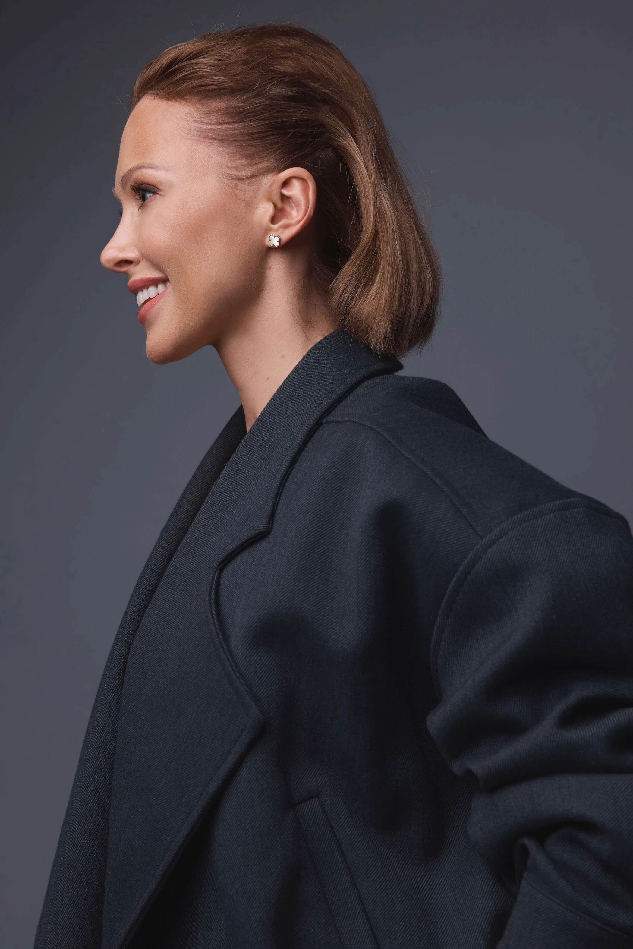 Side profile of a woman with short, light brown hair, smiling, wearing a black blazer and pearl earrings against a dark gray background.