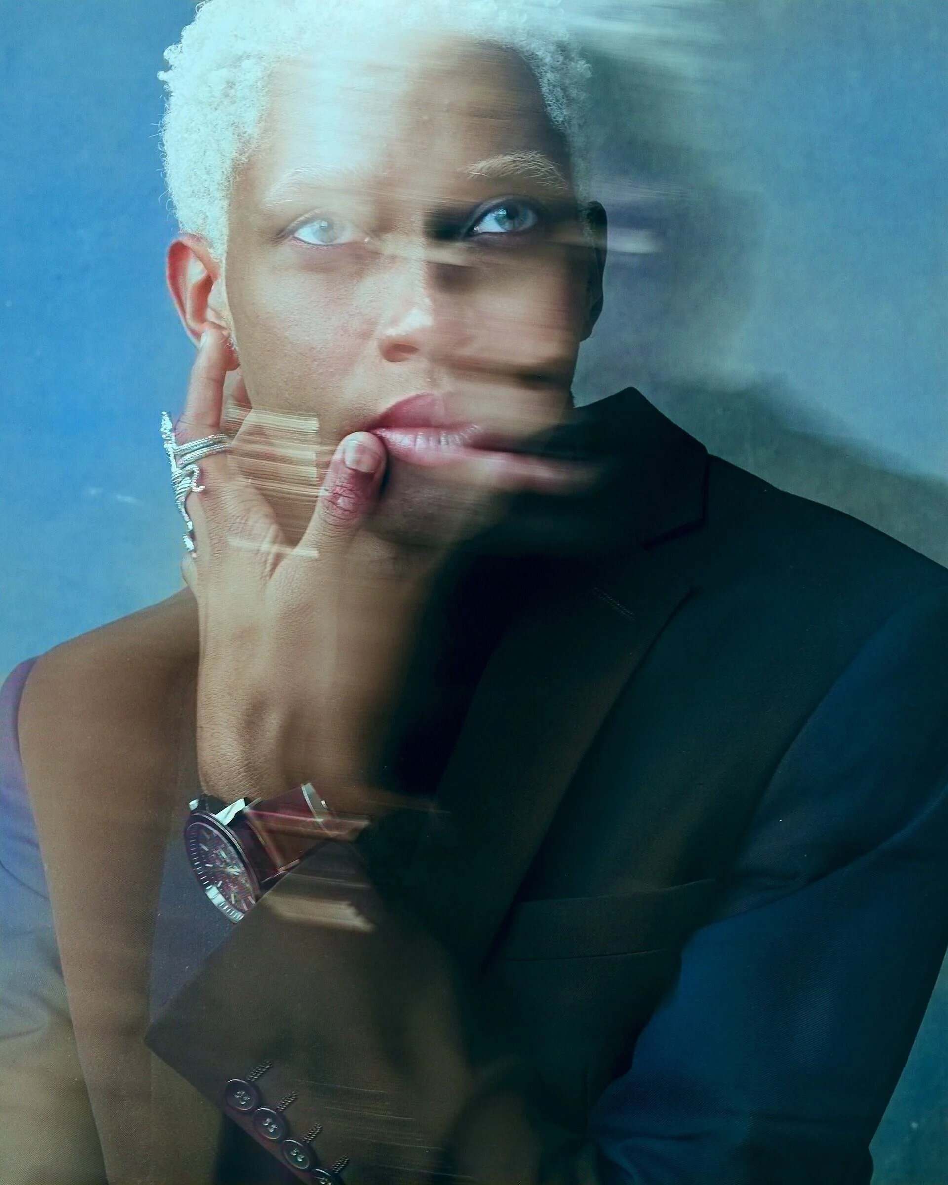 Person_with_short_white_hair_in_a_dark_suit_against_blue_backdrop.jpg