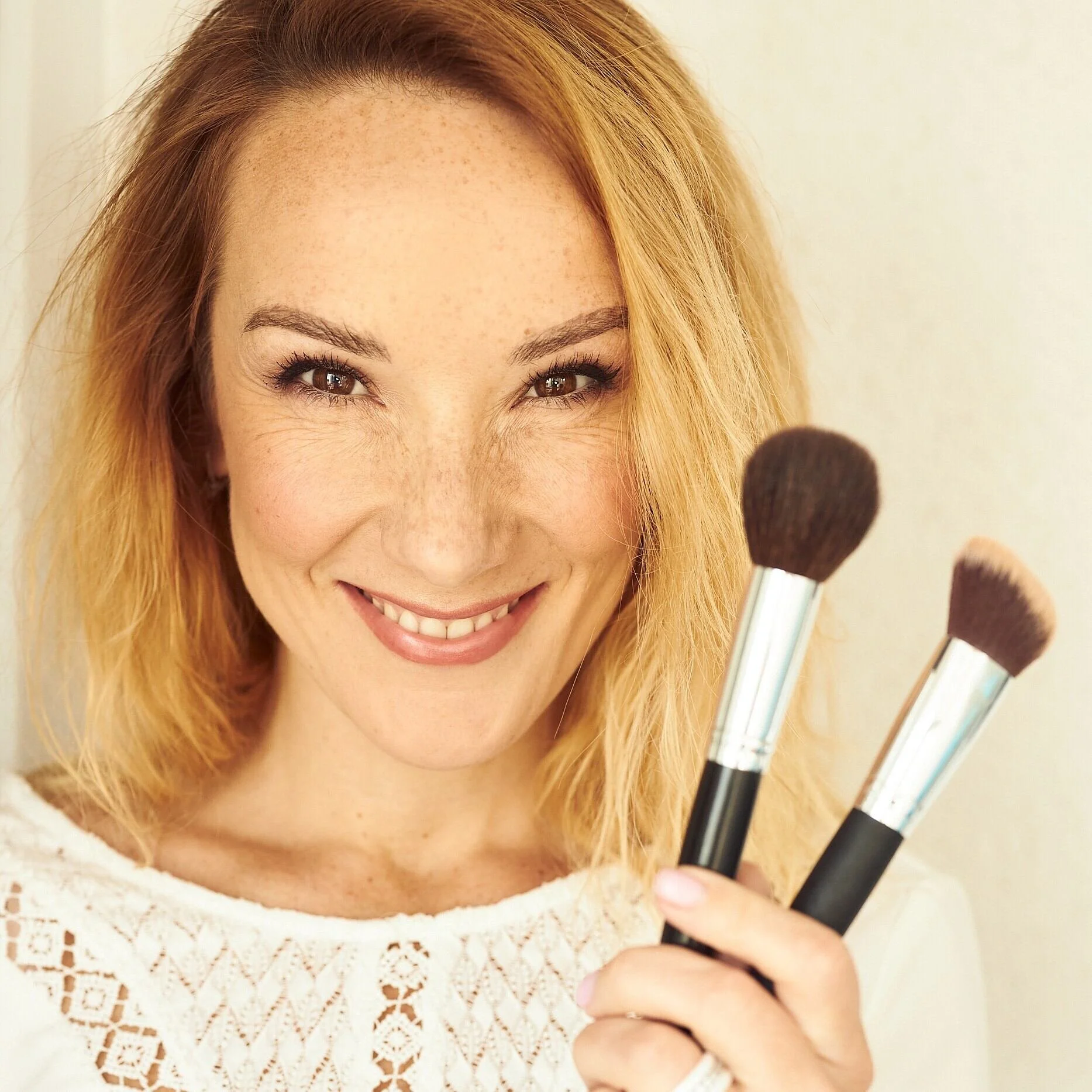A woman with blonde hair and freckles smiles while holding two makeup brushes. Captured in Geneva by a professional portrait photographer.