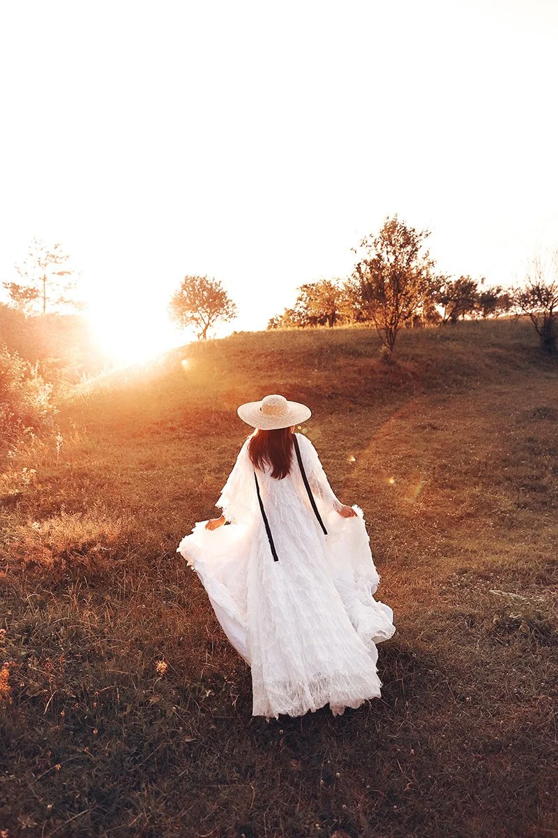 Femme_en_robe_blanche_et_chapeau_de_paille_marchant_dans_un_champ_au_coucher_du_soleil.jpg