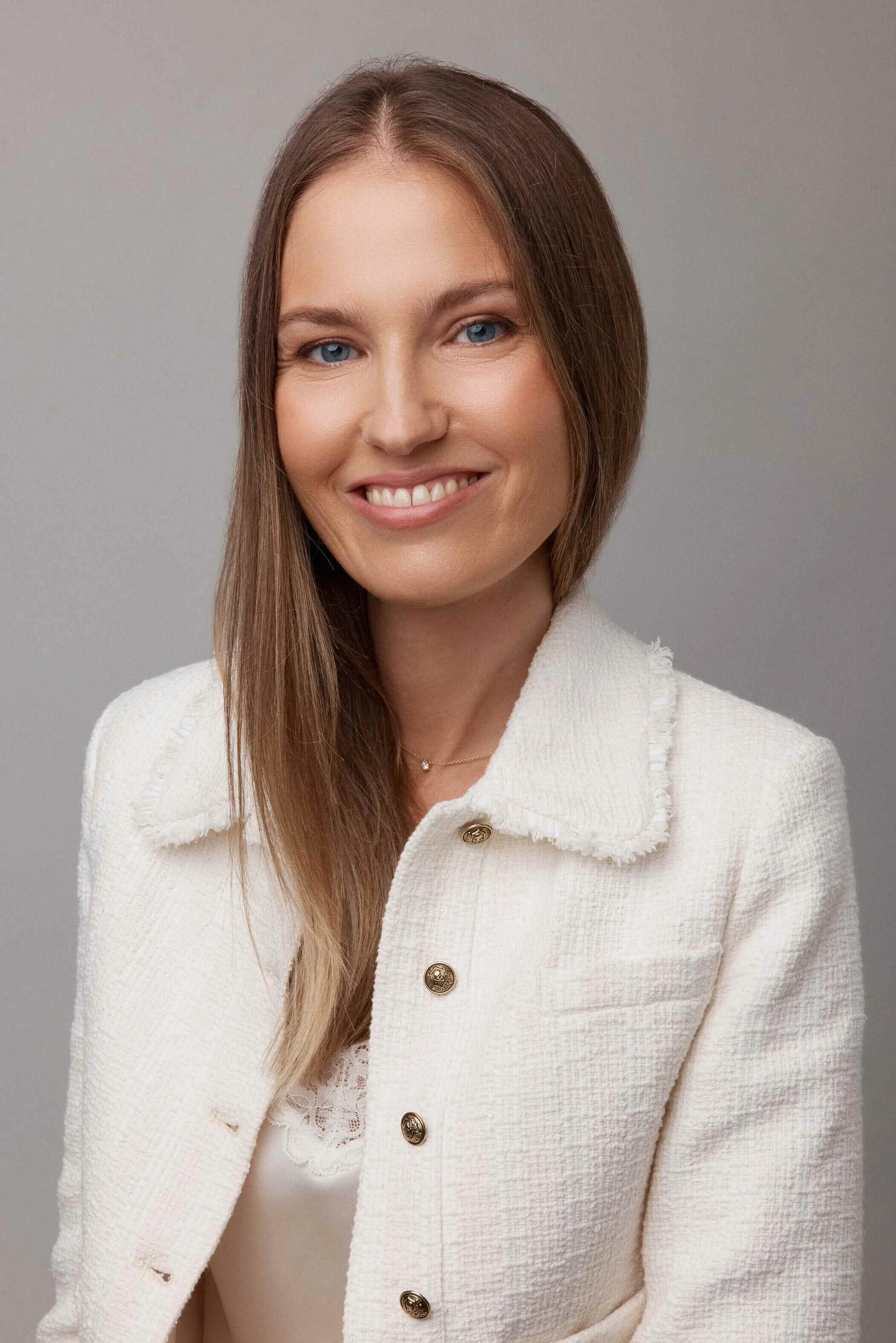A portrait of a woman with long light brown hair, blue eyes, and fair skin, smiling, wearing a white textured blazer with gold buttons and a delicate necklace, against a neutral gray background.