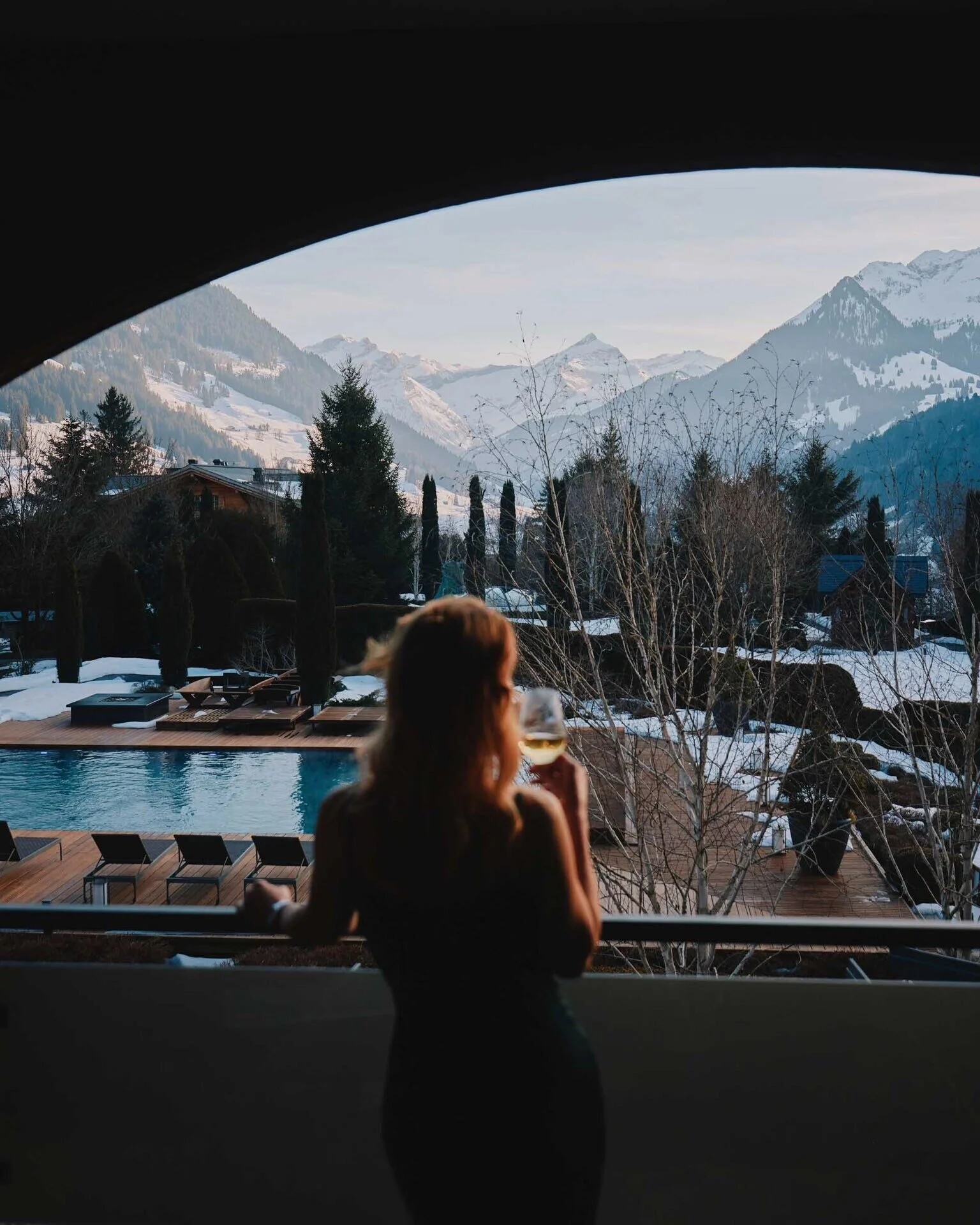 A woman in a black dress holds a glass while gazing at snow-capped mountains. Captured in Switzerland by a professional portrait photographer.