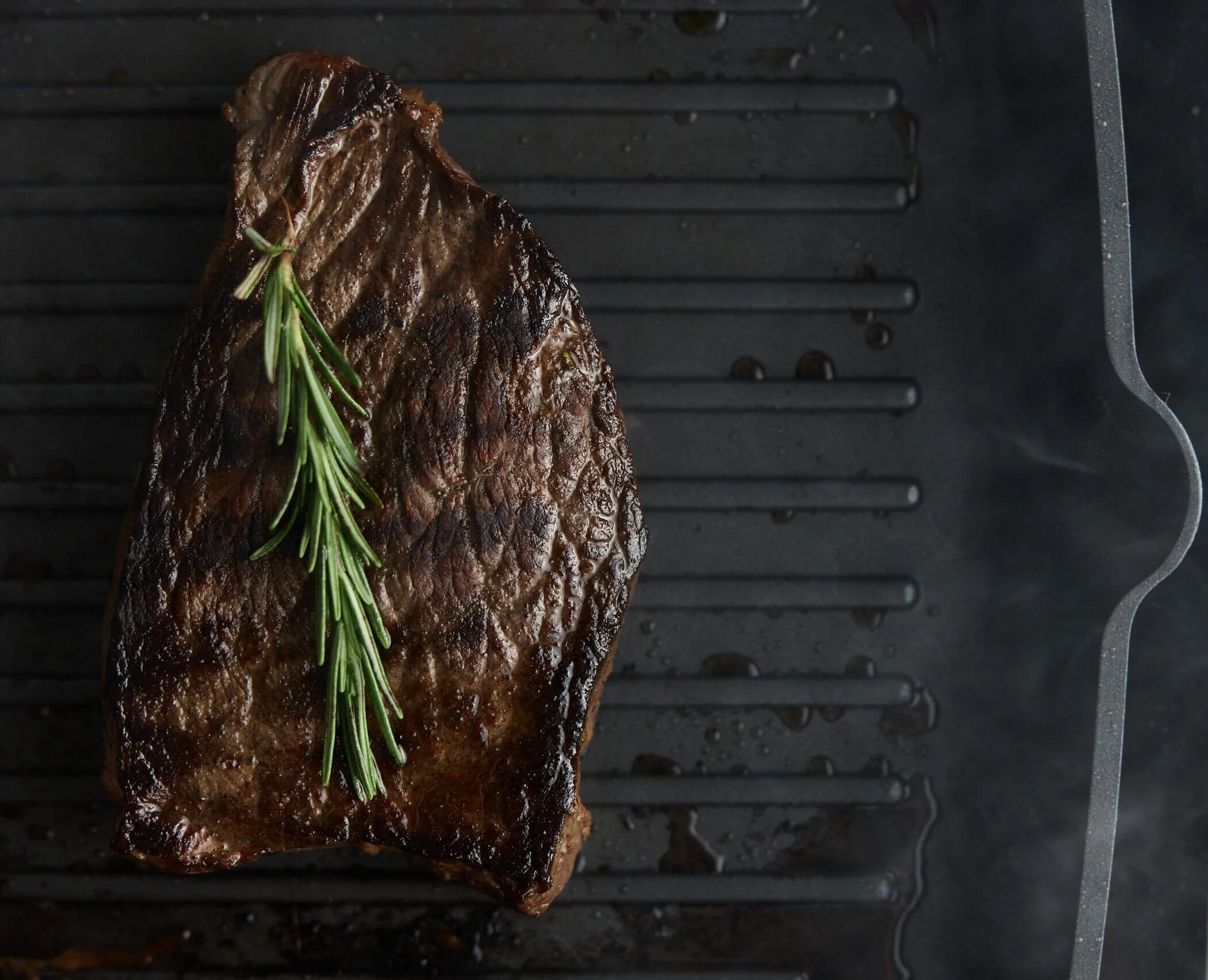 A grilled steak garnished with a sprig of rosemary sits on a black grill pan. Captured in Switzerland by a professional portrait photographer.