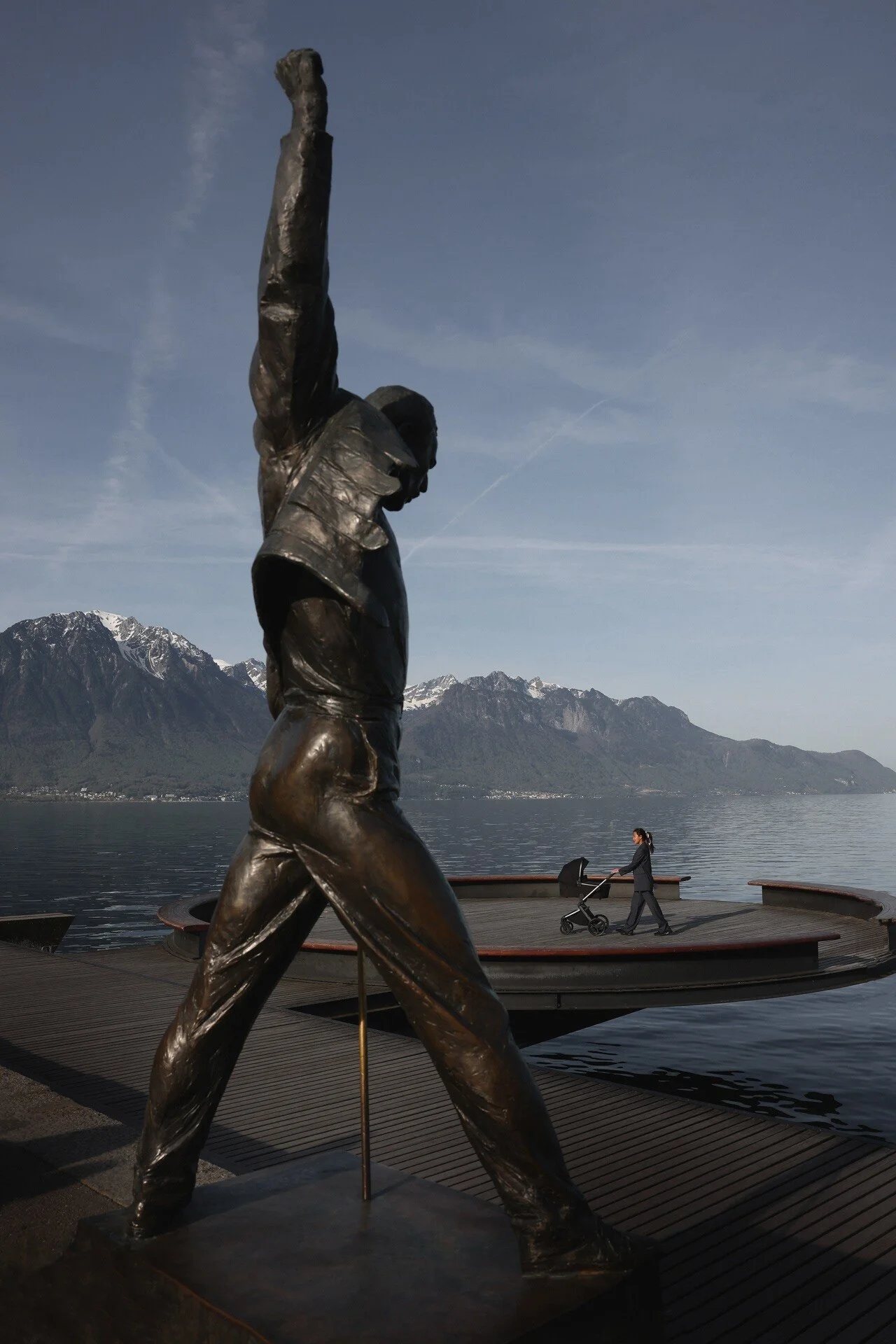 Une statue de bronze au bras levé se dresse près d'un lac, tandis qu'une femme promène une poussette au premier plan. Cette photo a été prise à Montreux par un photographe professionnel.