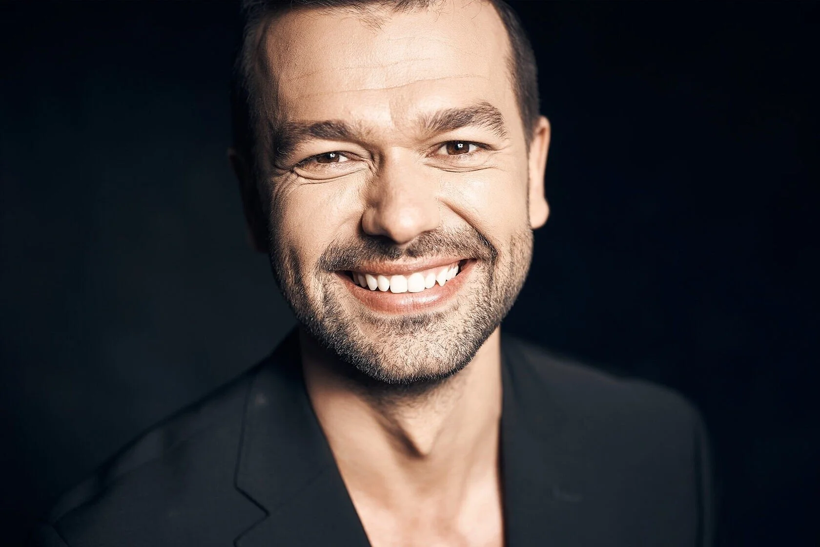 Smiling_man_in_black_suit_against_dark_background.jpeg